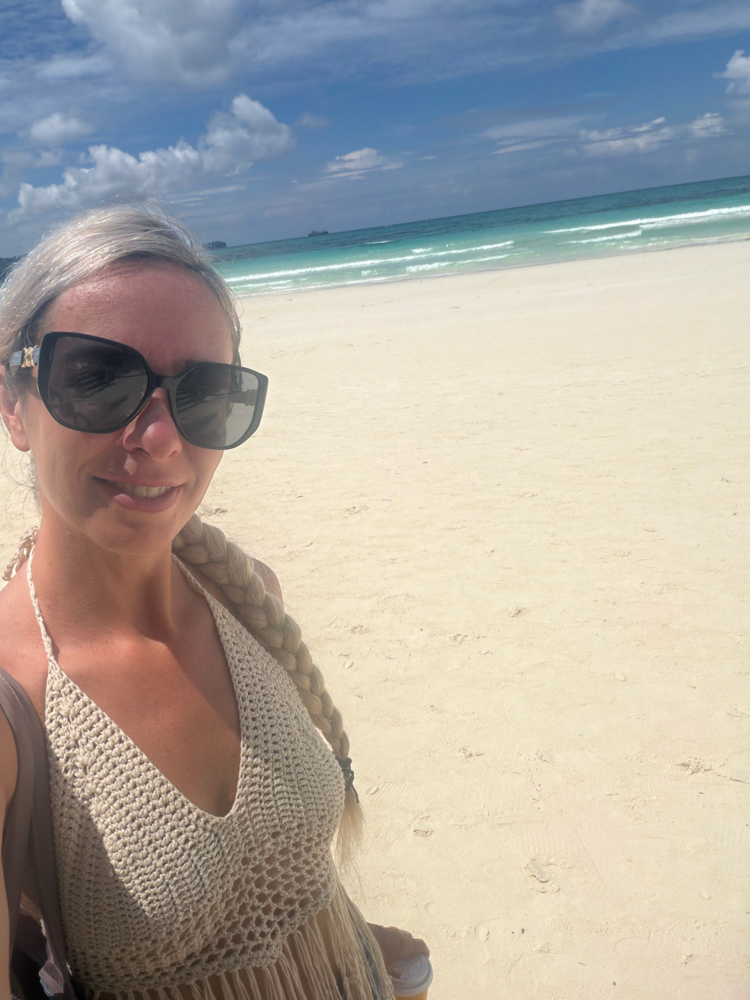 A woman with blonde hair and sunglasses takes a selfie on a bright, sunny day at a beach in Malay, Philippines. She is wearing a crochet halter top and has a braid of hair over her shoulder. The background shows a sandy beach, turquoise water with gentle waves, and a blue sky with scattered clouds. A ship is visible in the distance on the ocean. The overall scene depicts a relaxed tropical vacation.