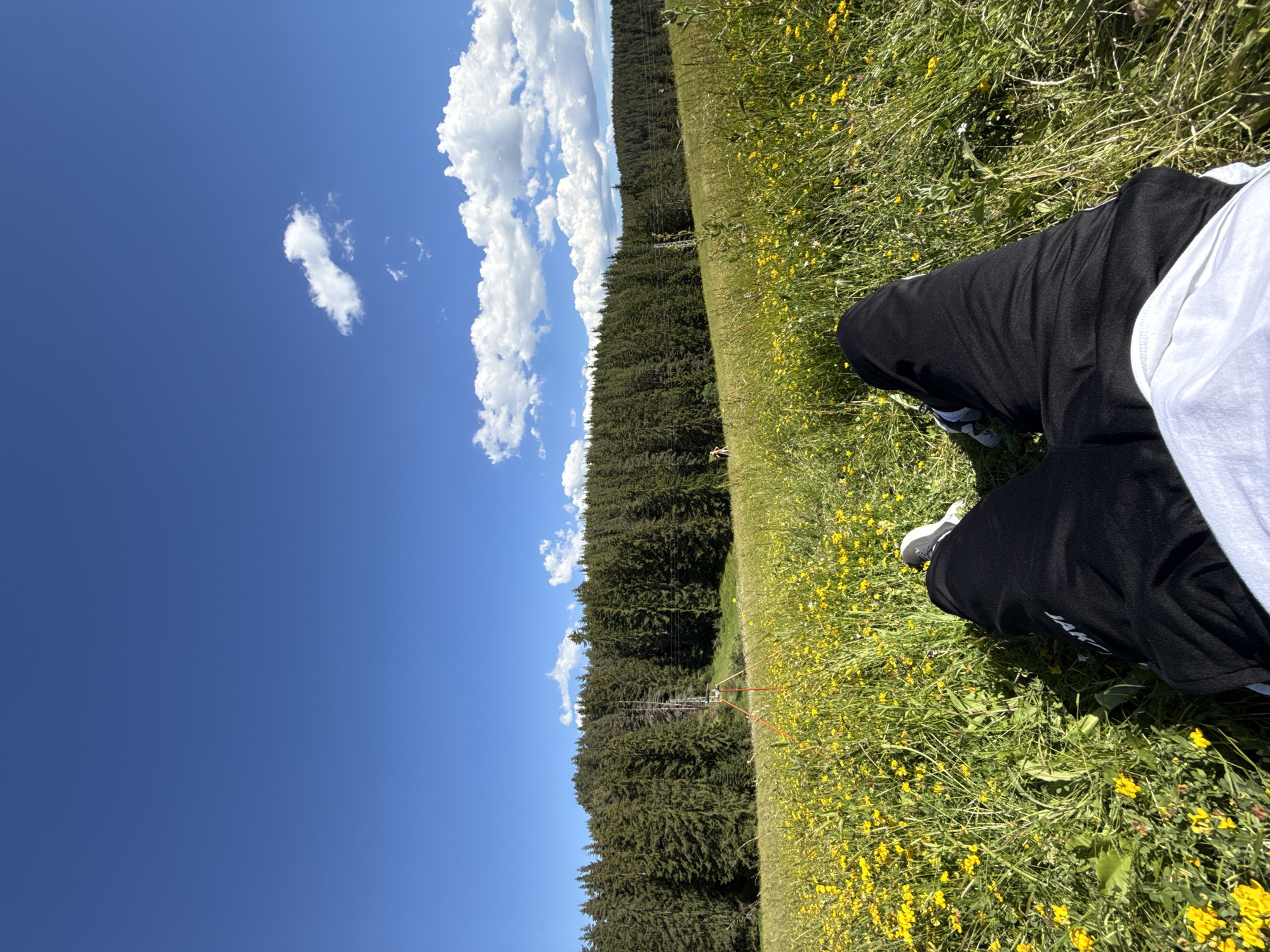 A first-person perspective shot captures a person's legs and feet as they recline in a meadow filled with yellow wildflowers. The meadow is on a gentle slope leading up to a dense evergreen forest. The sky above is a clear, bright blue with a few scattered white clouds. In the mid-ground, a tall, slender object, possibly a surveying tripod or a weather station, stands in the field with orange supports. Further in the background, a figure is visible walking in the meadow. The scene appears to be set in the Bulgarian mountain resort of Yundola, known for its natural beauty and forested landscapes. The lighting suggests it is daytime, likely mid-morning or afternoon, with the sun casting subtle shadows.