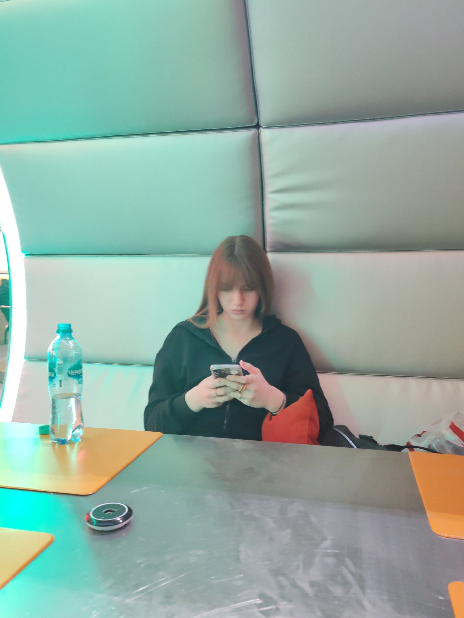 The image shows a young woman sitting alone in a modern, brightly lit indoor setting, likely a café or food court in Saint Petersburg, Russia. She has shoulder-length brown hair with bangs and is wearing a black hooded top. Her head is tilted downwards as she is engrossed in her smartphone, held with both hands.
She is seated in a booth with light-colored, padded backrests arranged in rectangular panels. A strong, artificial turquoise-green light emanates from the left side, casting a distinct hue over the booth and the table.
In front of her, on a reflective grey table, there is a clear plastic water bottle with a blue cap. Partially visible Cyrillic text on the bottle's label appears to be "ЧЕРНИ", possibly part of "Черноголовка" (Chernogolovka), a Russian beverage brand. Also on the table are two orange rectangular placemats or trays, and a small, black and silver circular device. An orange cushion is placed behind her on the bench, and a dark bag is visible further to her right. The woman appears focused on her phone, suggesting she is waiting, browsing, or texting. The overall atmosphere is cool and contemporary due to the lighting and decor.