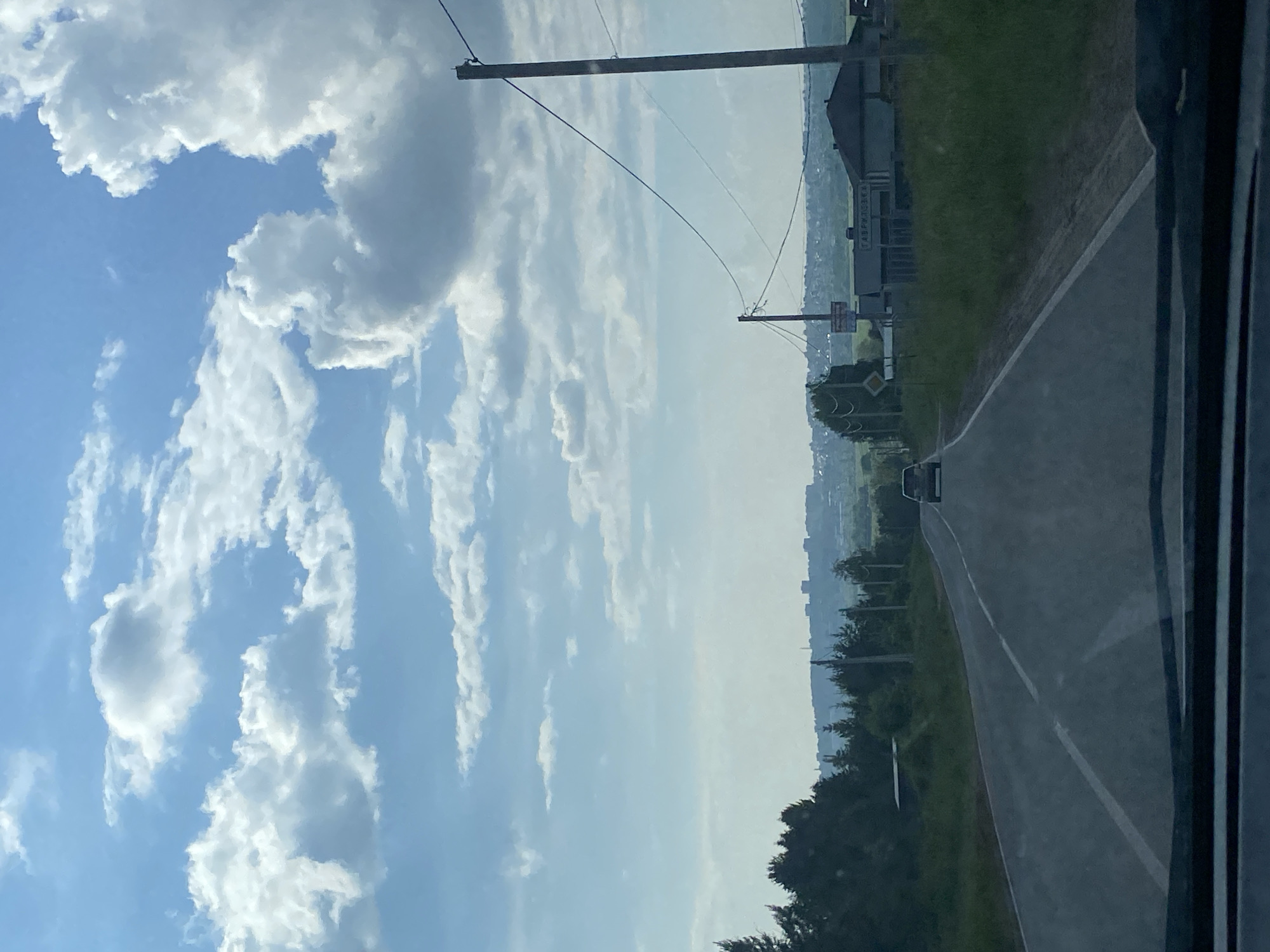 The image depicts a view from a vehicle traveling on a paved road in the city of Kamstskoye, Russia. The road has white lane markings and is flanked by a grassy verge and trees. In the distance, a city skyline is visible with buildings and a forest. The sky is blue with fluffy white clouds. There are utility poles with wires alongside the road. One pole has a sign attached to it. A single car is visible on the road ahead.