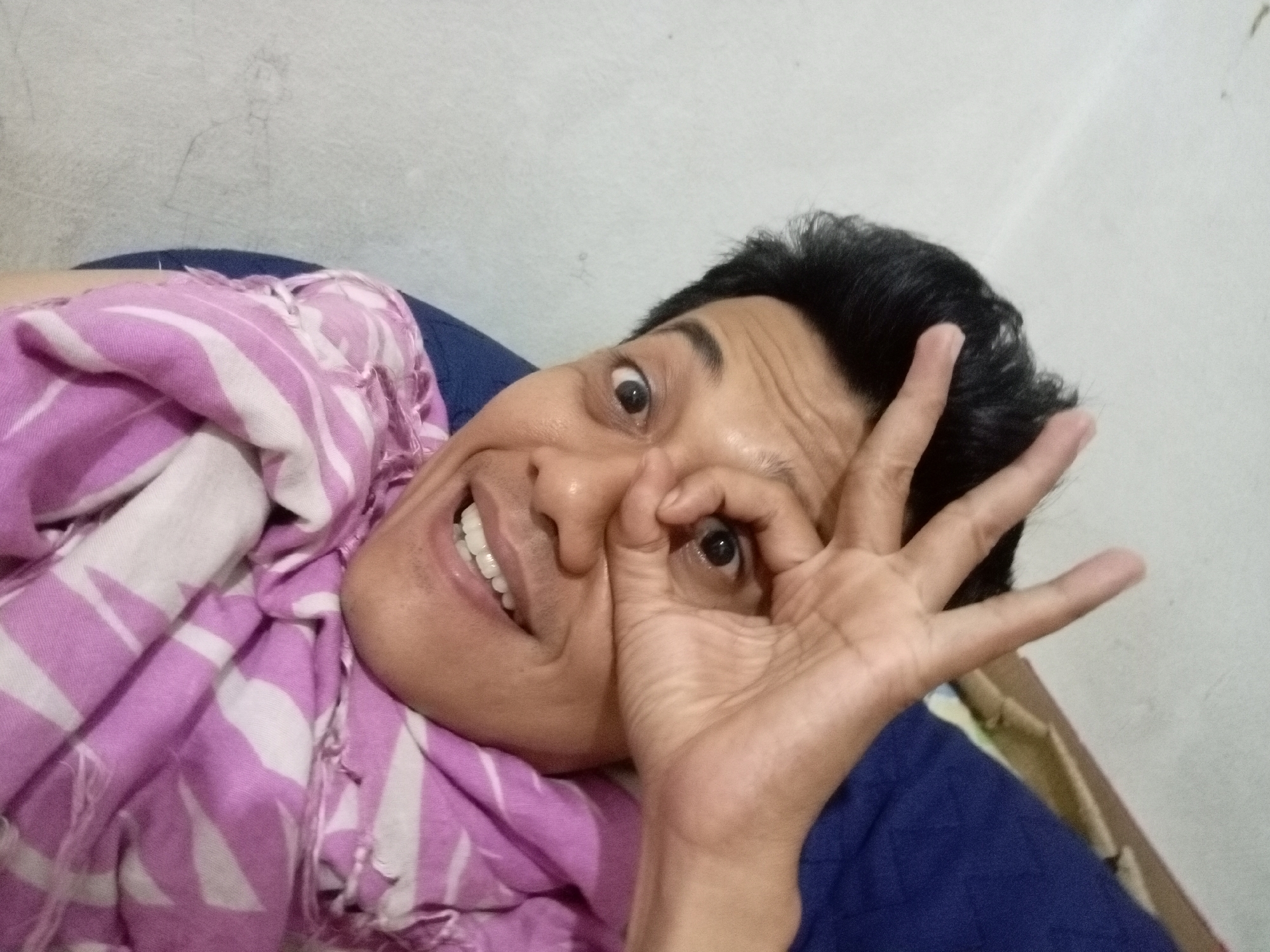A person is lying down, wrapped in a pink and white striped blanket, with their head resting on a blue surface. They are looking at the camera with their left eye covered by their right hand, forming a circle with their thumb and index finger. The person has dark hair and is smiling, showing their teeth. The background appears to be a white or light-colored wall. There is no specific information about the location in Batam, Indonesia that can be determined from the image itself.