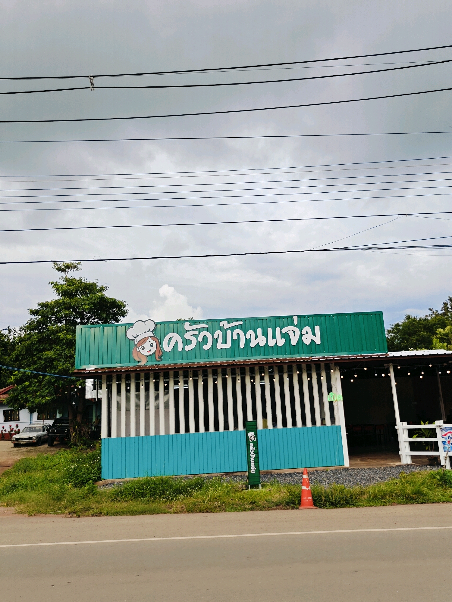 This is a daytime, eye-level, outdoor, vertical, medium shot photograph taken in Mu Si, Thailand. The foreground is dominated by a road with a white line. To the left of the road, there is a grassy area with some plants and a small, orange traffic cone. Behind the grassy area, there is a building with a green, corrugated metal roof. On the roof, there is a sign with a cartoon of a woman wearing a chef's hat and the Thai text "ครัวบ้านแจ่ม" (Krua Baan Jaem). The building has a light blue base and white vertical slats. In the background, there is a large tree, a white car, and a black SUV parked in front of a house. The sky is overcast with clouds.