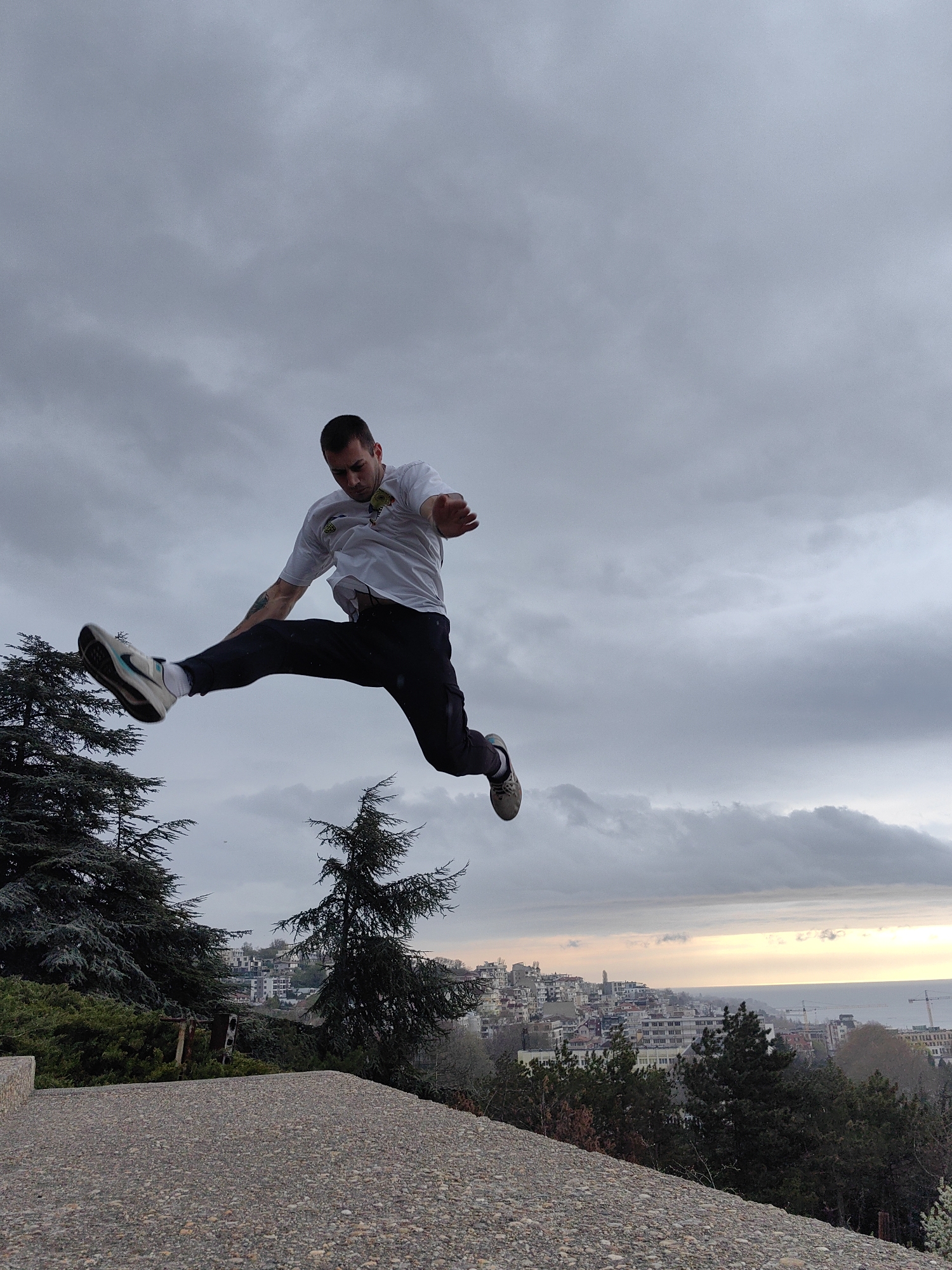 A man is captured mid-air in a dynamic jump, positioned against a backdrop of a cloudy sky. He is wearing a white t-shirt, dark pants, and sneakers. His body is angled, and limbs are outstretched. A tattoo is visible on his left arm.
Below him, there is a concrete surface, likely a rooftop or elevated platform. In the distance, there is an urban landscape featuring buildings, trees, and the sea. It is Varna, Bulgaria, as the architecture and the presence of the Black Sea coastline are typical to this city. The scene is likely in the Asparuhovo district, from the elevated perspective and view of the sea. There is a soft glow on the horizon, suggesting it may be sunset or sunrise.