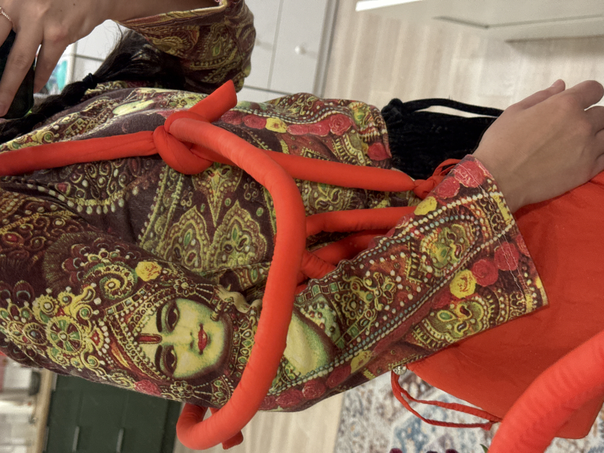 A person is shown from the shoulders up, wearing a brown patterned top with a face print on it and orange accessories. They are in an indoor environment. No information suggests a specific location like Krasnoyarsk, Russia.