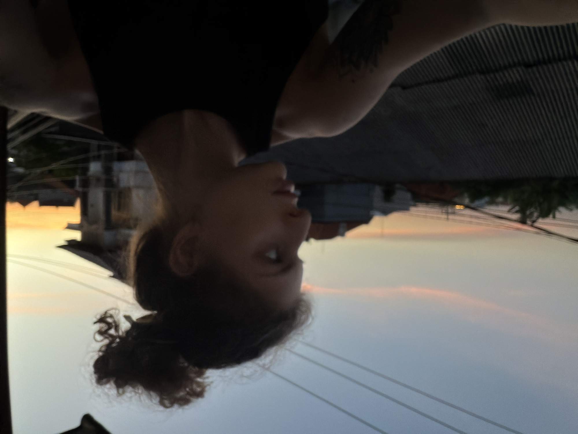 A low-angle, rotated photo shows a woman with a tattoo on her arm. She wears a black top and has her hair in a bun. The background depicts a city setting at sunset, including buildings, power lines, and vegetation. North Kuta, Indonesia is a possible location, based on the type of buildings and foliage.