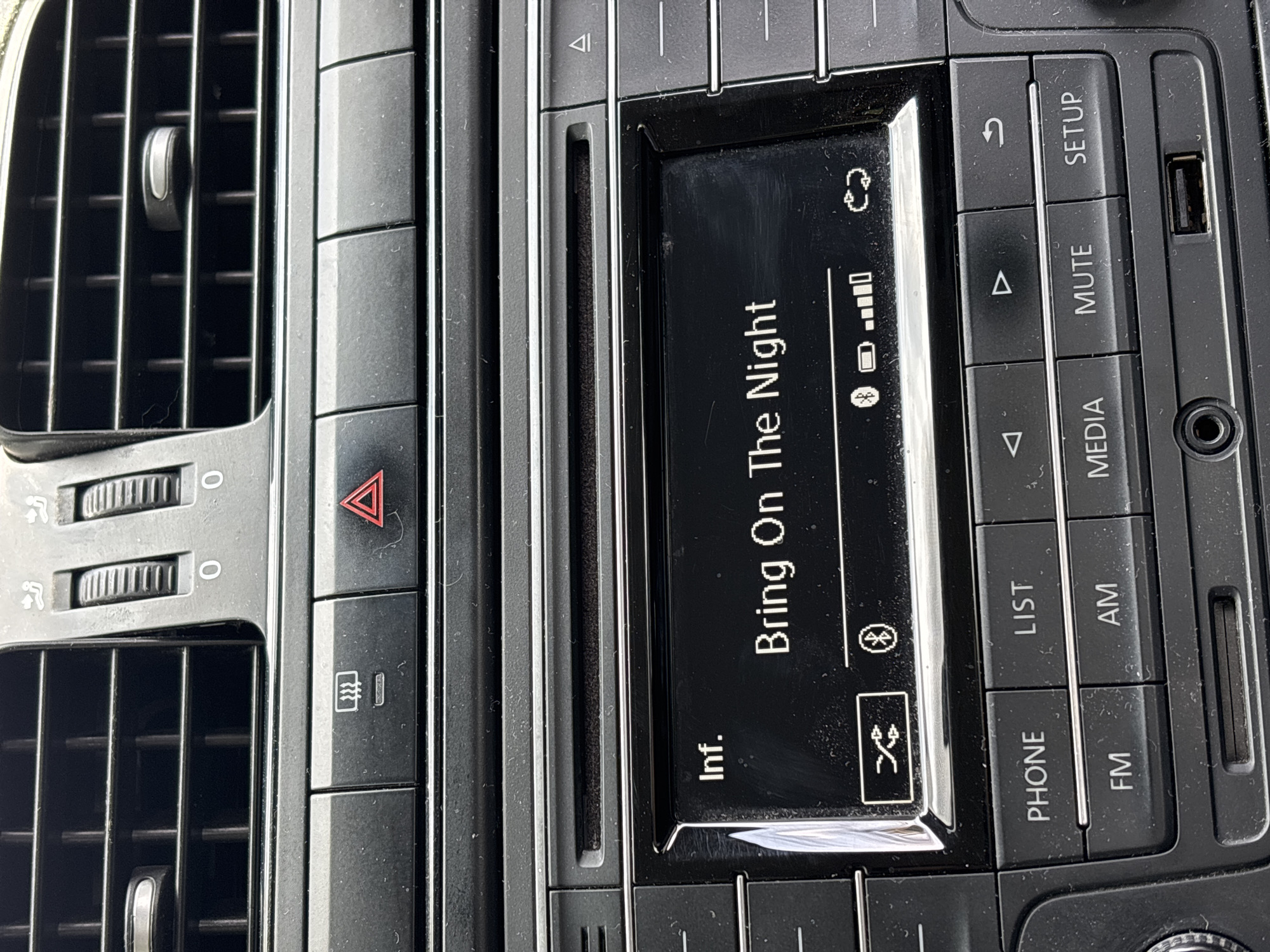 This media file captures the interior of a car, focusing on the central console area which includes air vents, climate controls, and the infotainment system. The setting is a car interior.
The main subject is the car's infotainment screen, which is displaying the text "Bring On The Night" and "Inf.", likely indicating a song title and information. Various icons are visible on the screen, including a refresh/sync symbol, a signal or battery strength indicator, a Bluetooth logo, and a shuffle icon, suggesting music is being played via a connected device. The screen itself appears slightly dusty.
Surrounding the screen are numerous control buttons. On the right side, buttons are labeled "SETUP", "MUTE", "MEDIA", "AM", "FM", "PHONE", and "LIST". There are also up and down arrow buttons, and a back/return arrow symbol. A USB port and an auxiliary audio input jack are visible on the far right of the console. To the left of the infotainment system are two rotary knobs, each marked "0", likely for air vent control, and a button with a defrost/recirculation symbol. Above these is a prominent red triangular button for hazard lights. The air vents themselves are visible on the far left.
The lighting suggests it is daytime, and the overall impression is of a standard vehicle dashboard. The provided context indicates the location is Puebla, Mexico.