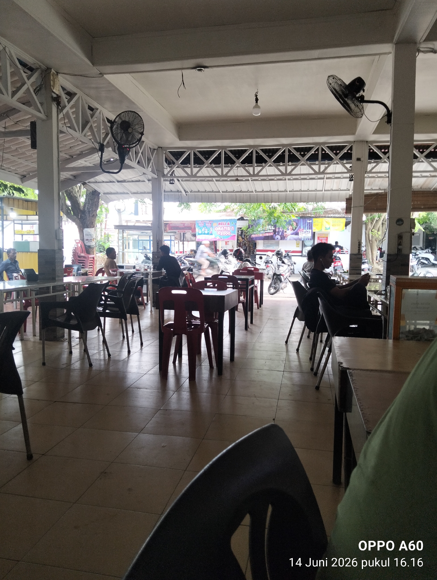 The image captures an interior view of an outdoor seating area, likely a cafe or restaurant, with tables and chairs arranged on a tiled floor. Natural light filters in from an open-air front, revealing a street scene with motorcycles and pedestrians. In the foreground, black plastic chairs and tables are visible, some with red chairs at other tables. Several ceiling fans are mounted on the structure, and a light bulb hangs from the ceiling. In the background, people are seated at tables, and a motorcycle is being ridden past the establishment. The scene is located in Lhokseumawe, Indonesia, as indicated by the timestamp "14 Juni 2026 pukul 16.16" which translates to "June 14, 2026 at 4:16 PM." The overall impression is of a casual, possibly a roadside eatery, in a tropical climate.