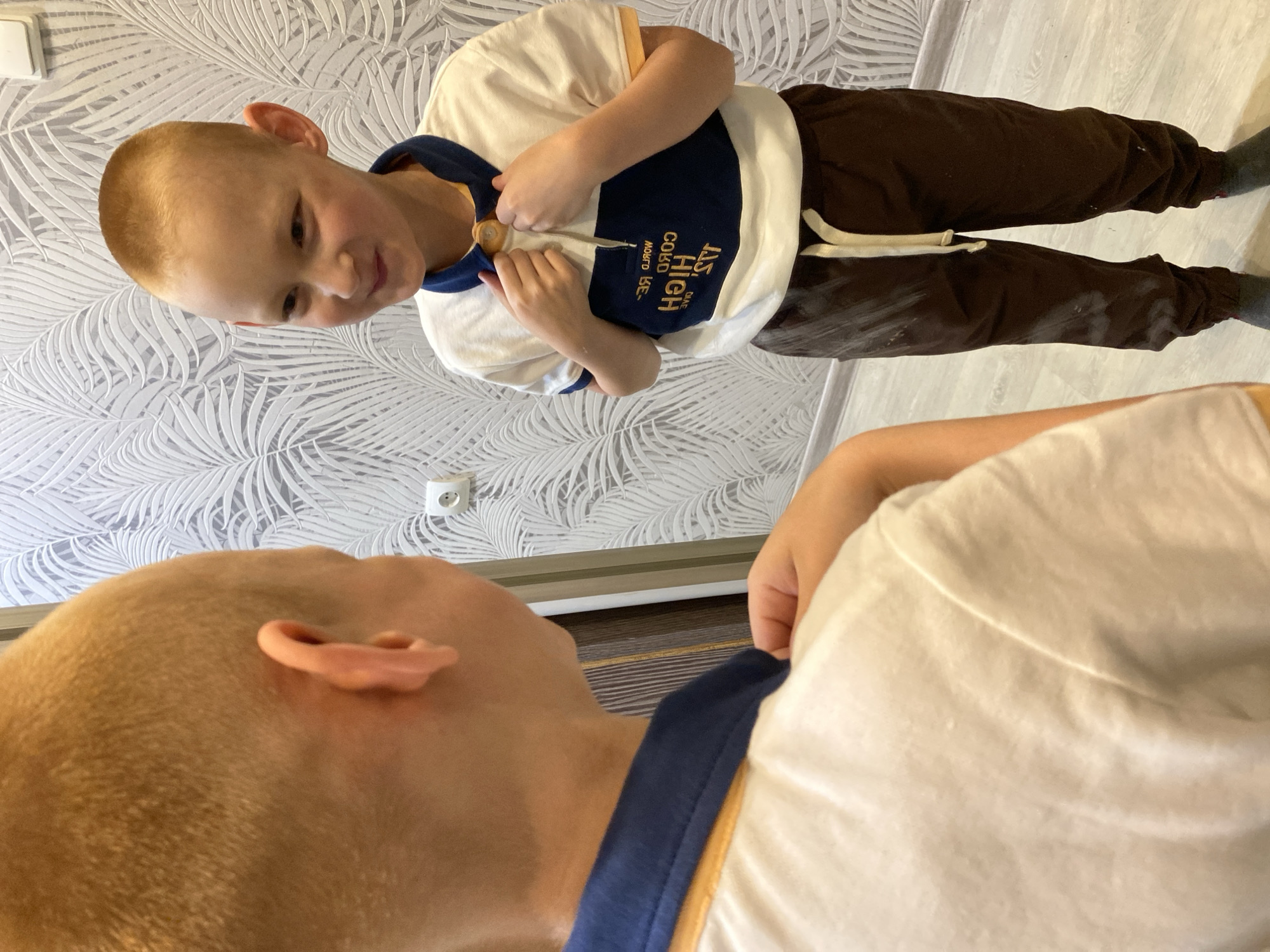 A fair-skinned boy with a shaved head is standing in front of a mirror. He is wearing a white t-shirt with navy blue accents and brown pants with a white drawstring. The boy is looking at himself in the mirror and adjusting the button on his shirt. The environment includes a light-colored wooden floor and a wallpaper with a leaf pattern. The location is Noviy, Russia, based on text visible on the boy's shirt.