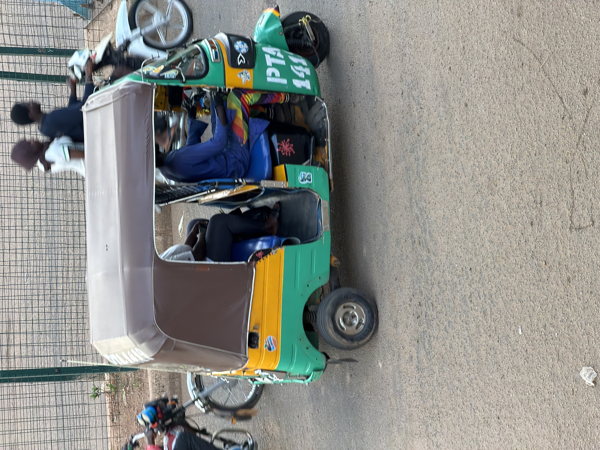 In Bukuru, Nigeria, a vibrant three-wheeled auto-rickshaw, painted in green and yellow with a brown canopy, is parked on a paved surface. The auto-rickshaw appears to be in use, with several people inside. One individual, wearing a blue outfit, is seated in the passenger area. In the foreground, two individuals are visible, one wearing a dark shirt and the other a white shirt with a green stripe on the collar. Another person, wearing a purple head covering, is also partially visible. A white motorcycle with a white rim is parked behind the auto-rickshaw. The scene suggests a typical day in Nigeria, with the auto-rickshaw likely used for local transportation. The lighting indicates it is daytime. The words "PTA 141" are prominently displayed in white on the green side of the auto-rickshaw.
