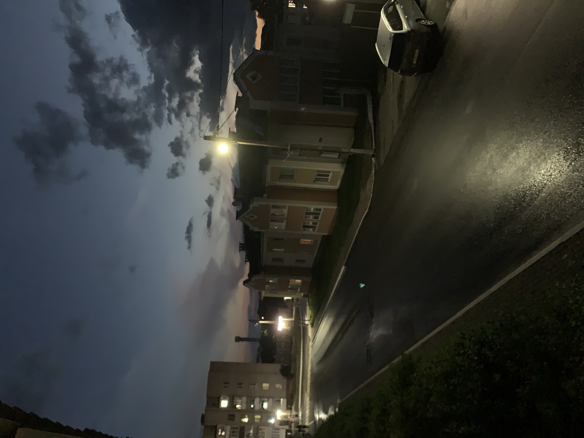 The image depicts a dimly lit urban street scene in Kapyĺ, Belarus, during twilight. On the left, a tall, multi-story apartment building with illuminated windows is visible. Adjacent to it, a wet asphalt road curves towards the right. On the right side of the road, a row of colorful, European-style residential buildings with peaked roofs and lit windows lines the street. A solitary, silver station wagon is parked on the right side of the road, its taillights dimly visible. A street lamp on the right illuminates a portion of the road and sidewalk. The sky is a deep blue, transitioning to hues of pink and orange near the horizon, with dark, dramatic clouds scattered across it. The wet road surface reflects the light, indicating recent rain. Green foliage is present along the left edge of the road. A distant tower-like structure can be seen on the horizon.