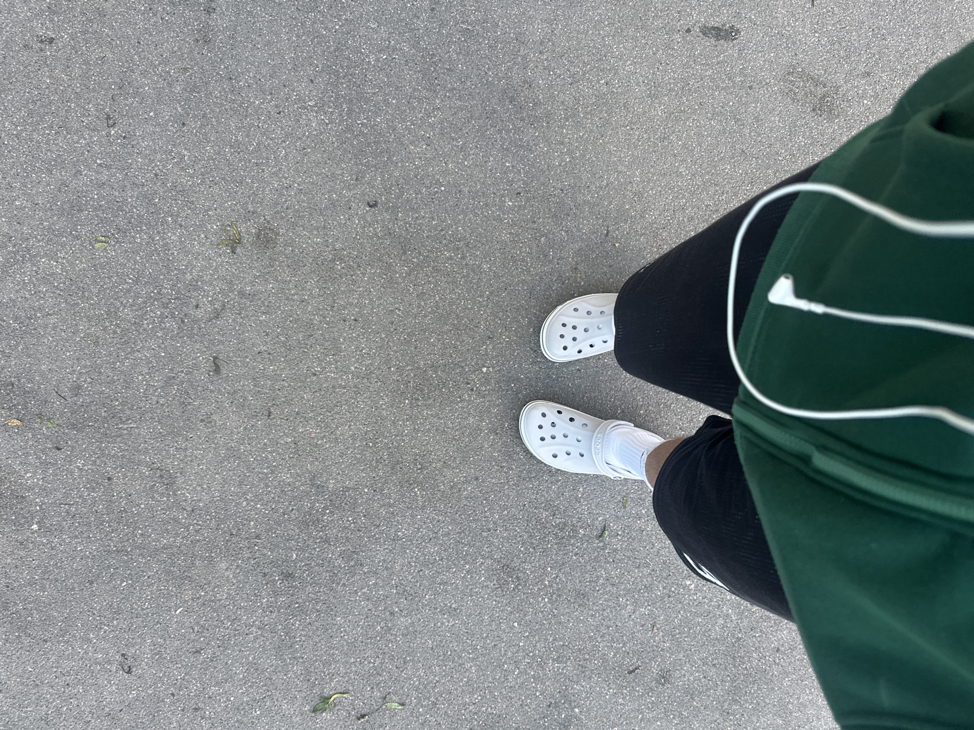 An overhead shot shows a person's legs and feet from the knees down, walking on a grey asphalt surface, possibly a sidewalk or path. The person is wearing white Crocs on both feet, white socks, black shorts, and a dark green hooded jacket. A white earbud cord is visible, draped across the jacket. Several small fallen leaves are scattered on the asphalt. The lighting suggests it is daytime.