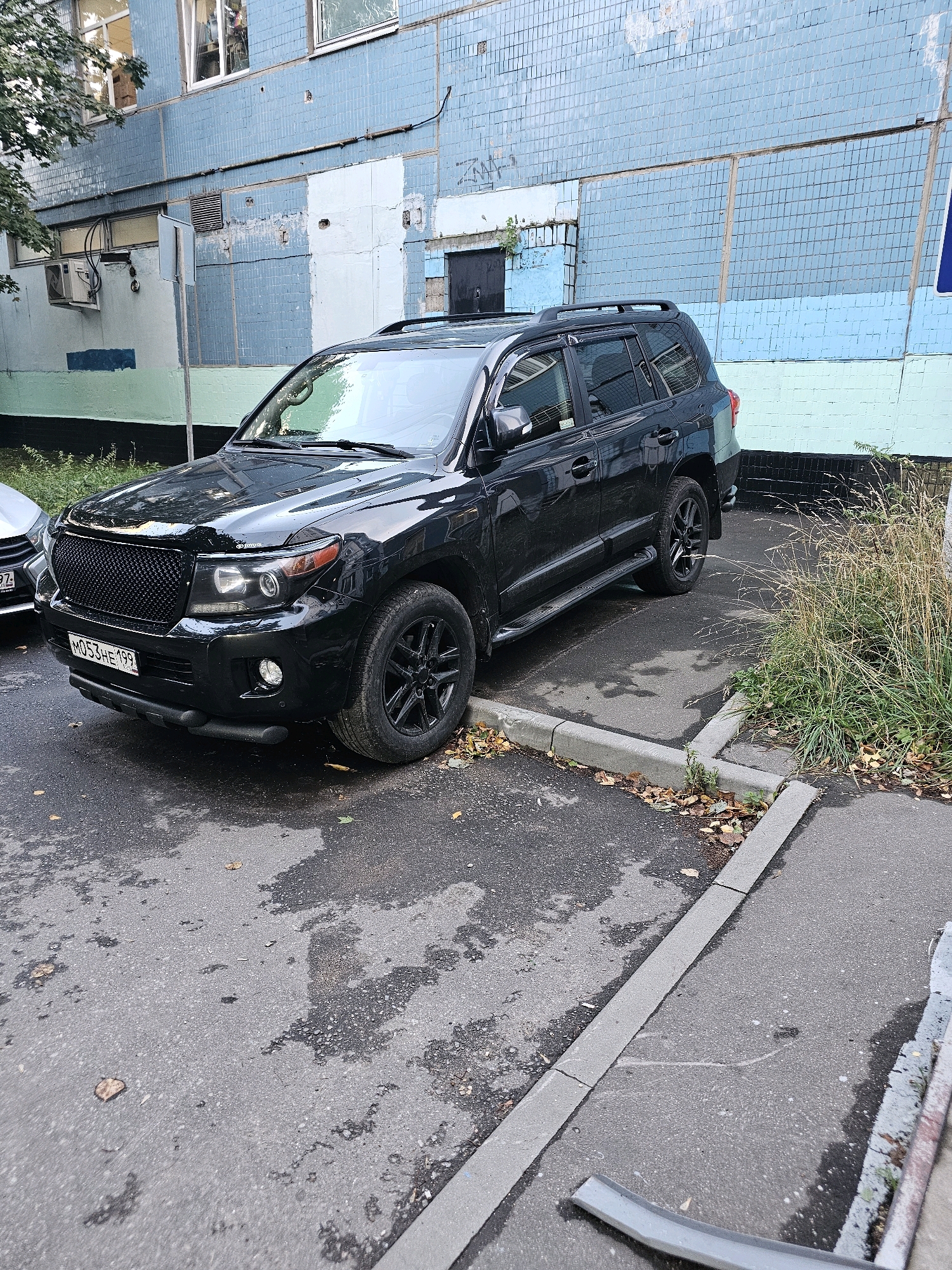 A black Toyota Land Cruiser is parked on a wet asphalt surface next to a light blue apartment building in the city of Khimki, Russia. The vehicle has a black grille and black wheels. The building's facade is partially tiled with light blue tiles and has some white patching. A tree with green foliage is visible on the left side of the image, and dry grass and fallen leaves are present near the building and along the curb. The license plate on the car is partially visible and appears to be Russian. The overall scene depicts a residential urban environment on a possibly overcast day.