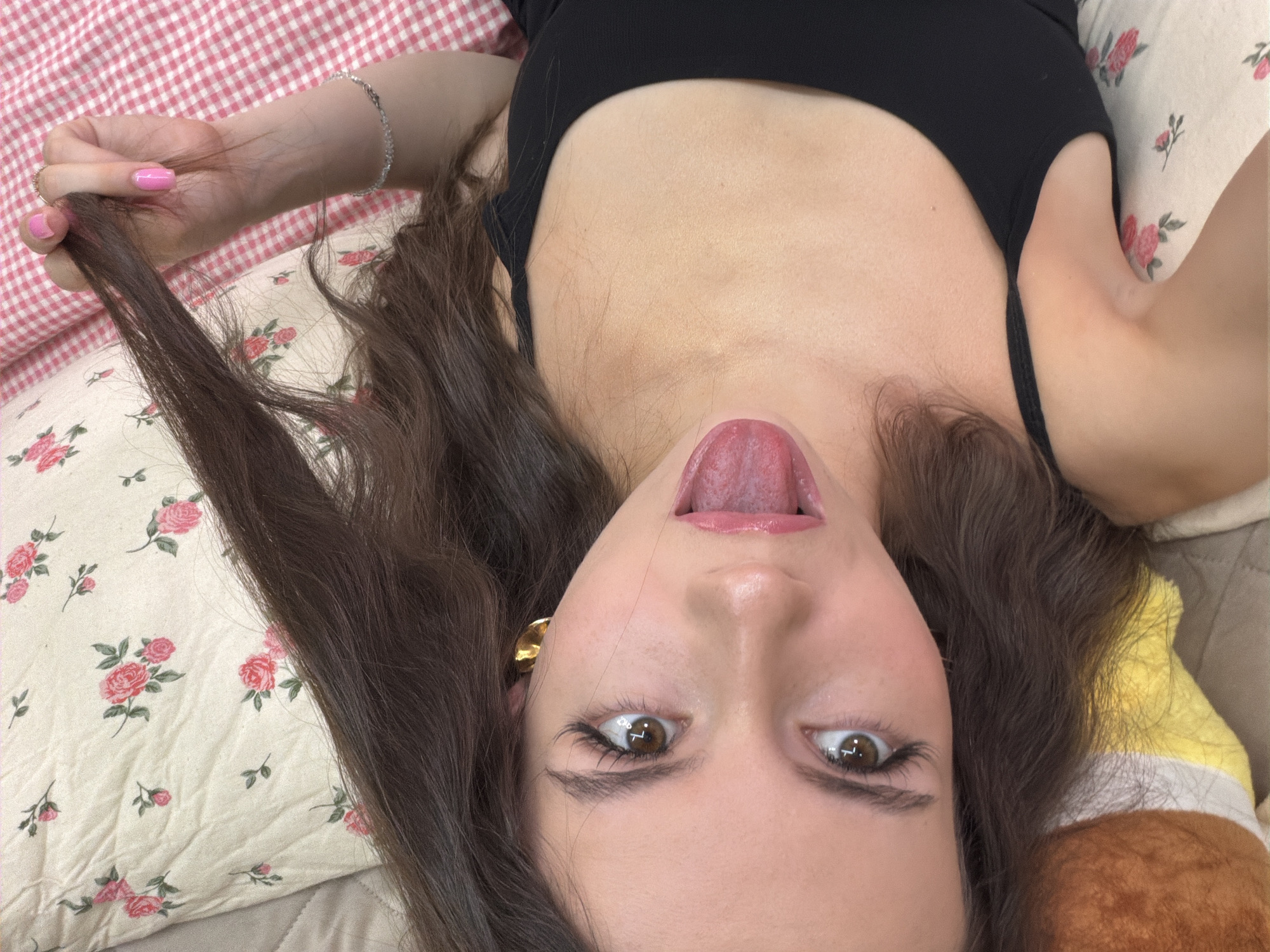 The image shows a young woman lying on a bed. She has long, dark hair and is wearing a black tank top. Her eyes are open and she is sticking her tongue out. She is wearing a gold earring in her left ear and a silver bracelet on her right wrist. She is also wearing a ring on her right hand. The bed is covered with a floral-patterned sheet and a pink and white checkered blanket. The image was taken in Chaikovsky, Russia.