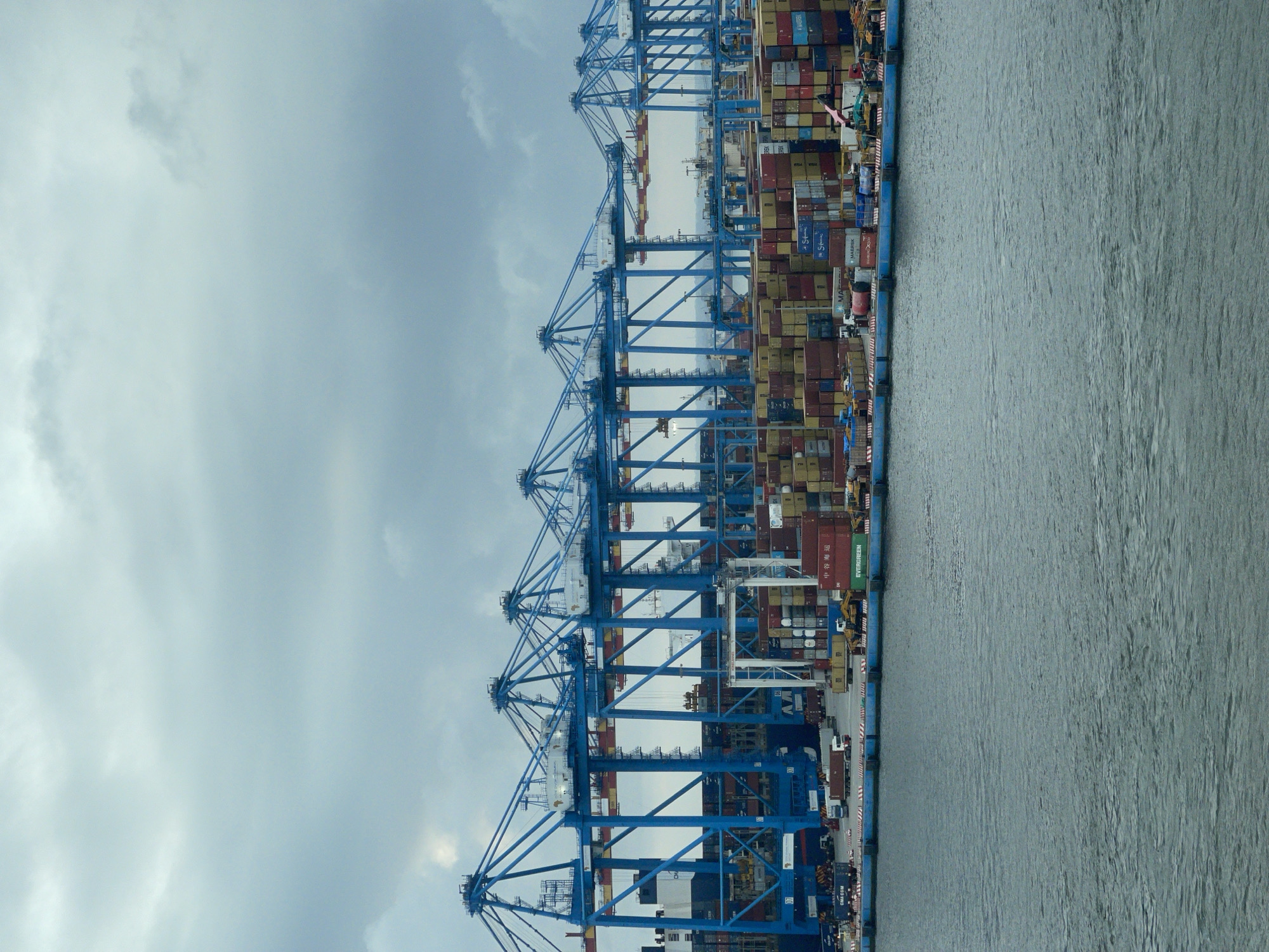 The image depicts a view of the Port of Abidjan, Ivory Coast. The foreground shows a body of water with ripples. The midground features a bustling container terminal with numerous shipping containers stacked high. Large, blue gantry cranes, characteristic of a port, dominate the skyline, suggesting ongoing operations. Several trucks and other port equipment are visible amongst the containers, indicating activity. In the background, a large cargo ship is docked. The sky is overcast and cloudy, contributing to a diffused light. The scene is devoid of any visible people, focusing solely on the industrial infrastructure of the port.