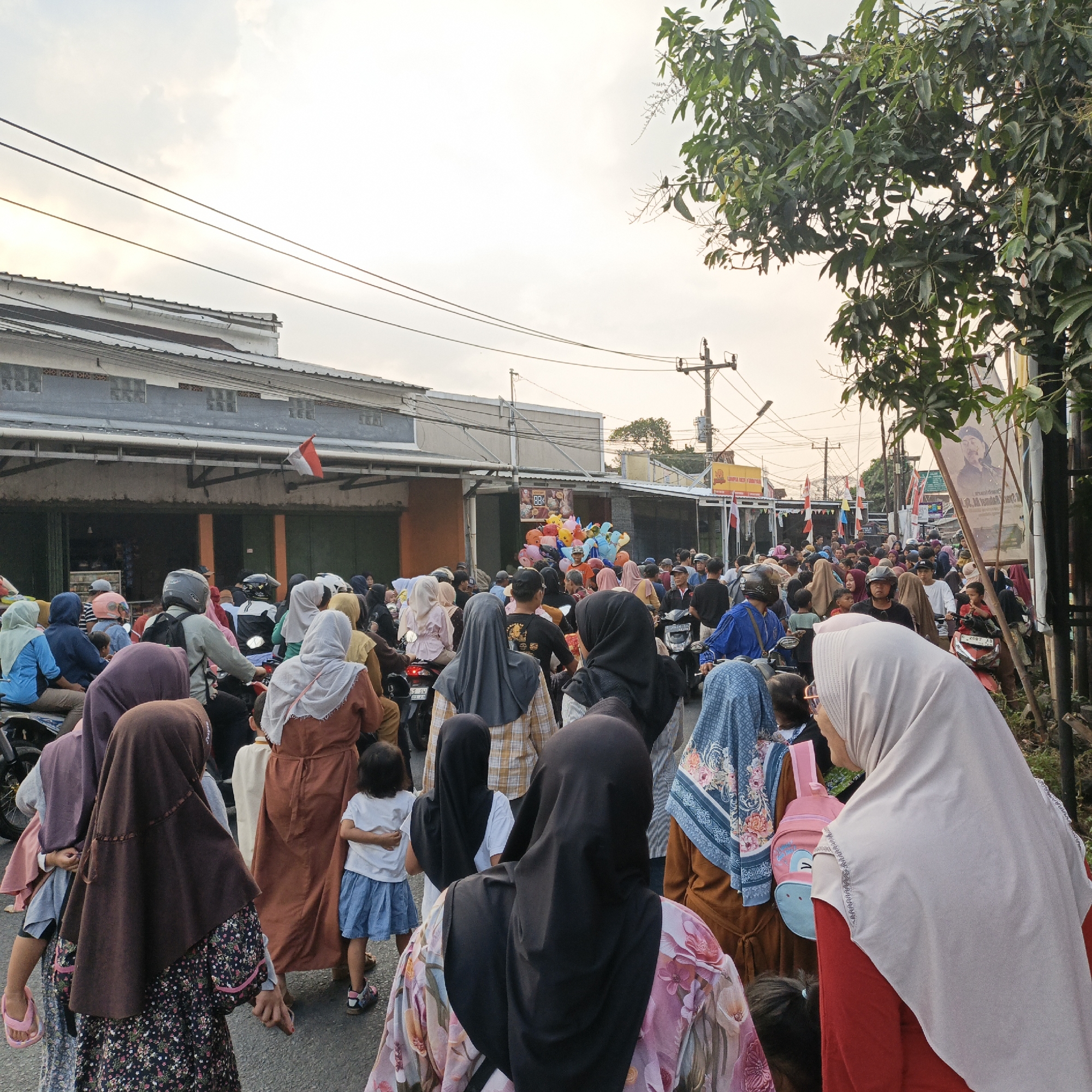 The image depicts a street scene in Randudongkal, Indonesia, filled with people, many of whom are wearing hijabs. The street is lined with buildings and appears to be a marketplace or a busy thoroughfare. A large crowd of people are walking, and some are riding motorcycles, suggesting a lively event or daily activity. Several motorcycles are visible, some with riders and passengers. Colorful balloons are clustered together, possibly for sale or decoration. Indonesian flags are also present, indicating a local celebration or national event. In the background, there are utility poles with electrical wires. The lighting suggests it is daytime, possibly late afternoon due to the soft light. The overall impression is one of a bustling community gathering.