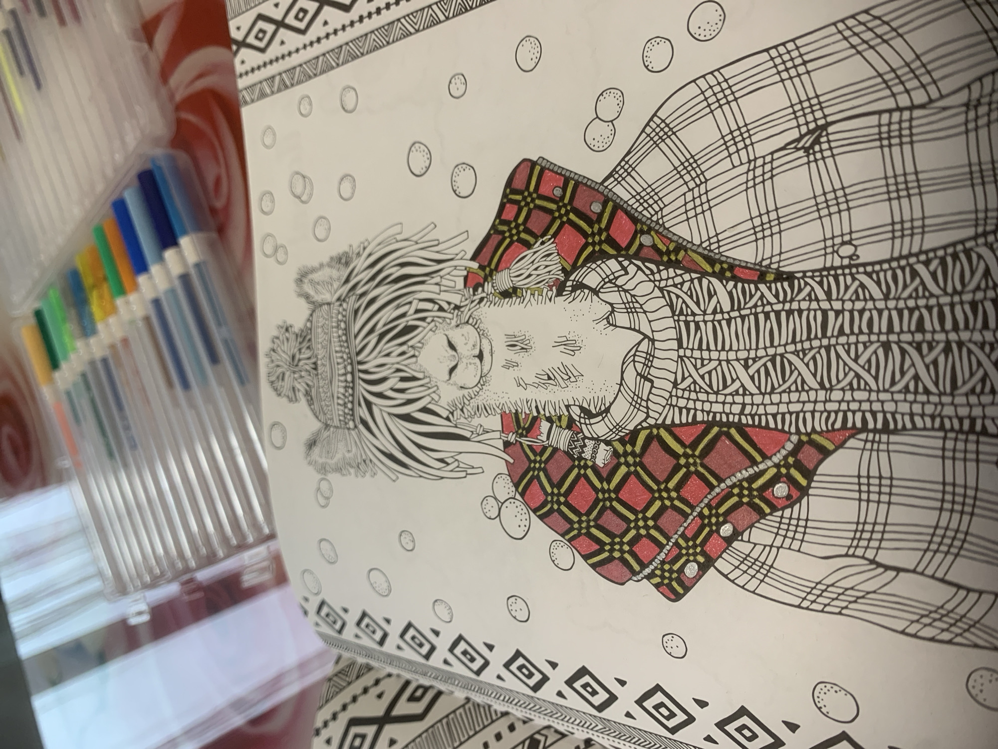 The image shows a partially colored-in illustration of a woman or girl lying down, wearing a hat and patterned clothing. The illustration is contained within a coloring book with patterned borders. To the left of the coloring book is a container of pens. There is no indication of the location of Izhevsk, Russia.