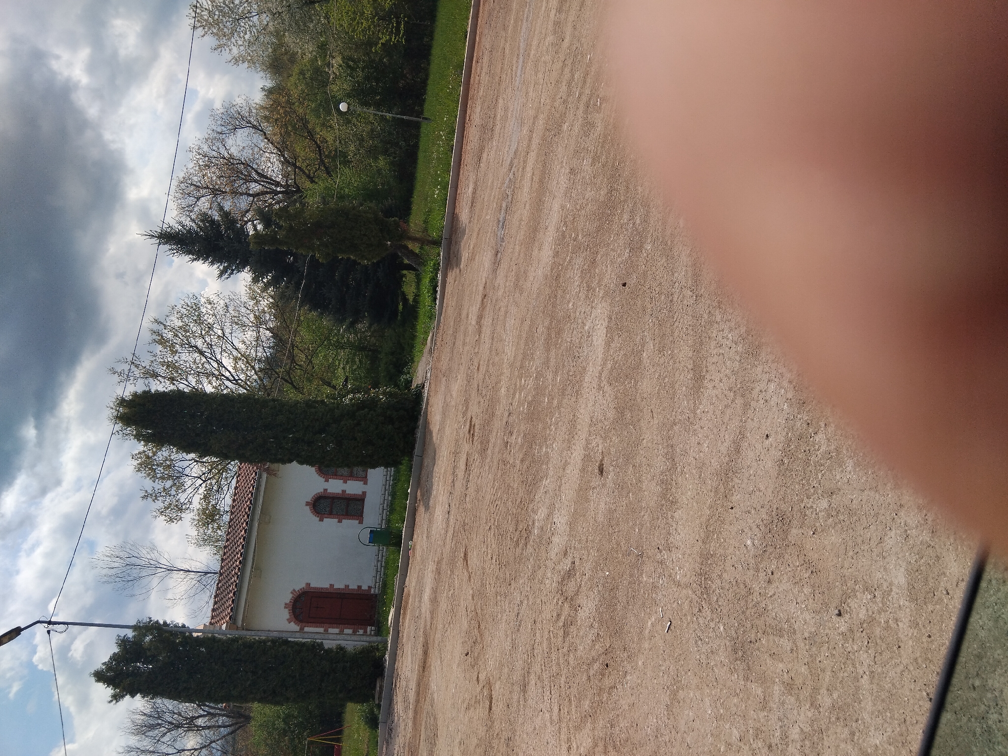 The image, taken in Velchevo, Bulgaria, presents an outdoor scene featuring a small building and various trees under a cloudy sky. A large, blurry, reddish-brown object, likely a finger or thumb, partially obstructs the view on the right side of the frame.
In the foreground, there is an unpaved, light brown, gravelly or dirt path. To the left, a small, white building with a traditional terracotta tiled roof is visible. It has two arched dark brown windows with white trim and a matching arched dark brown door. A green trash can is placed next to the building.
Several trees are present throughout the scene. Two tall, narrow, dark green coniferous trees stand prominently near the building. In the background, deciduous trees, some with bare branches and others with light green foliage, suggest either early spring or late autumn. A street lamp post with a light fixture is visible on the left side, and a thin wire or power line stretches across the sky.
The sky is largely overcast with dark grey clouds, though some patches of brighter sky are discernible. The scene is illuminated by natural daylight, indicating it was taken during the day. No people or specific activities are depicted; it is a static view of what appears to be a park, churchyard, or public area within the village. There is no visible text.