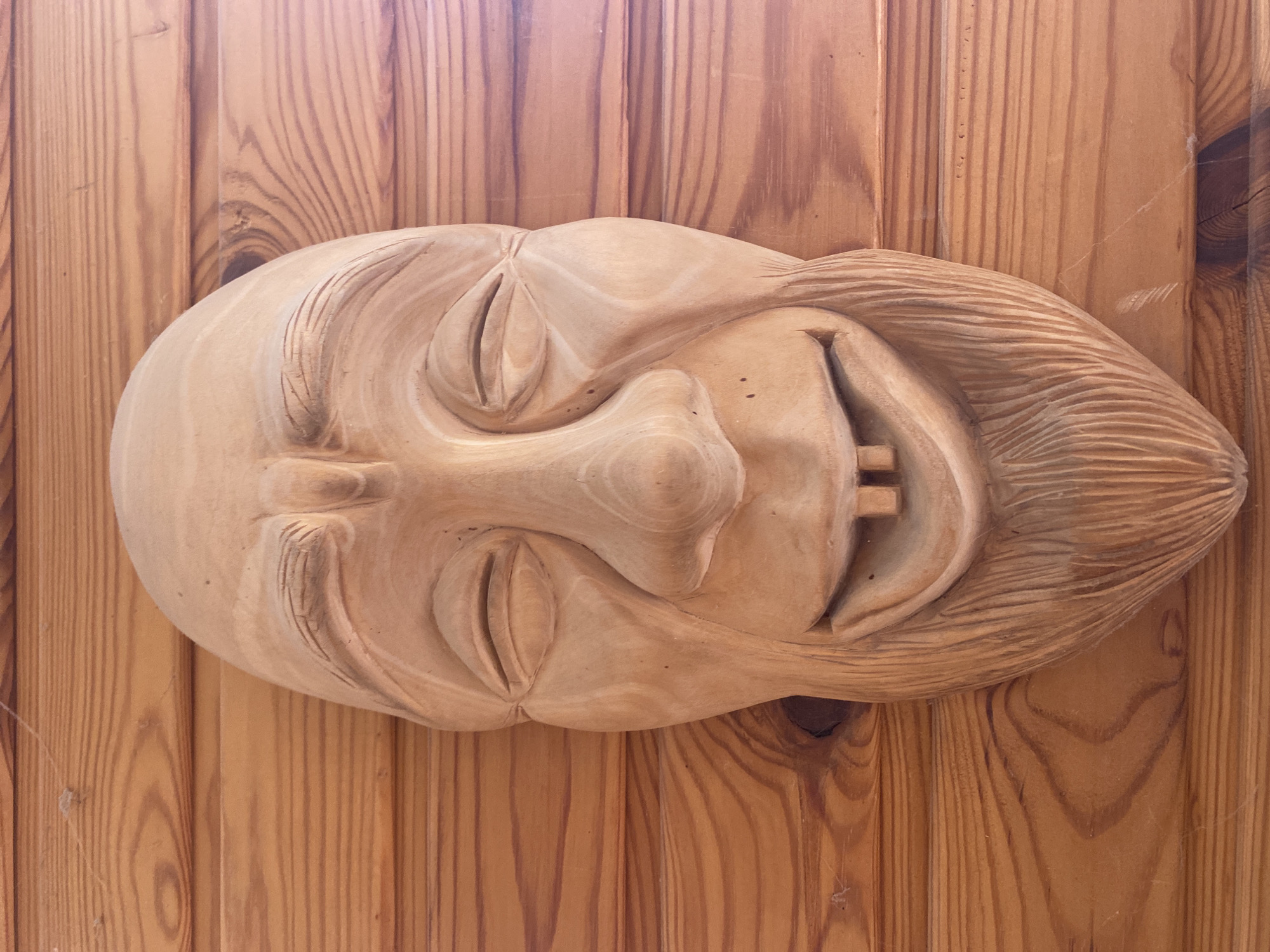 A carved wooden face is mounted on a wooden wall. The face has closed eyes, a large nose, and a smiling mouth with prominent teeth. The hair is also carved from wood. The wall is made of vertical wooden planks. The image was taken in Прикамский, Russia.