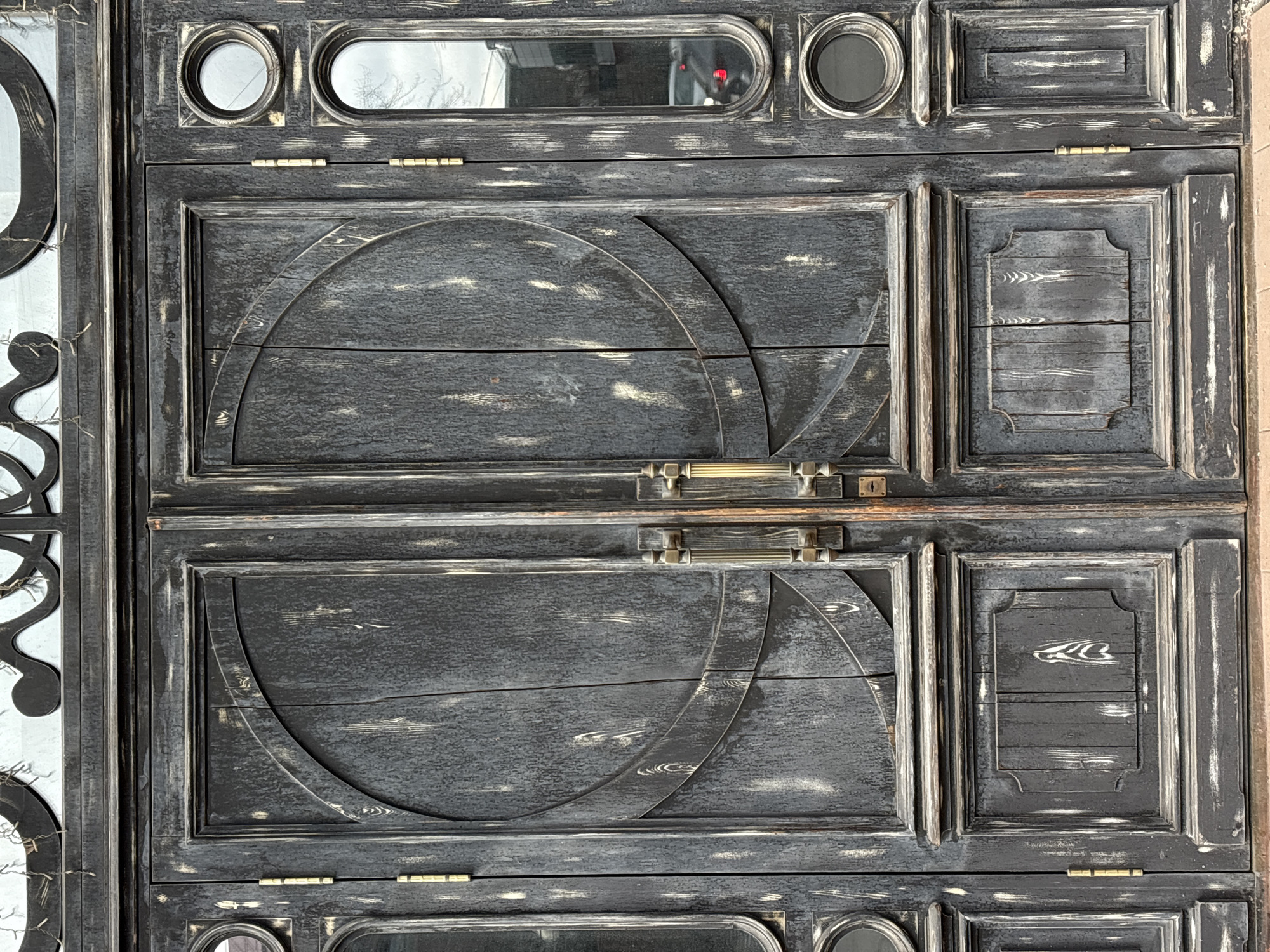 The image shows a close-up of a weathered, dark-painted wooden gate or door. The design incorporates geometric shapes, including circular arcs and rectangular panels. Some panels feature small, round or rectangular glass inserts. The door is fitted with a brass handle and hinges. The wood has a distressed finish, with white paint visible through the dark topcoat. There is no evidence of people or animals in the image. The location is unspecified, and the text is unable to infer a location in Krasnoyarsk, Russia.
