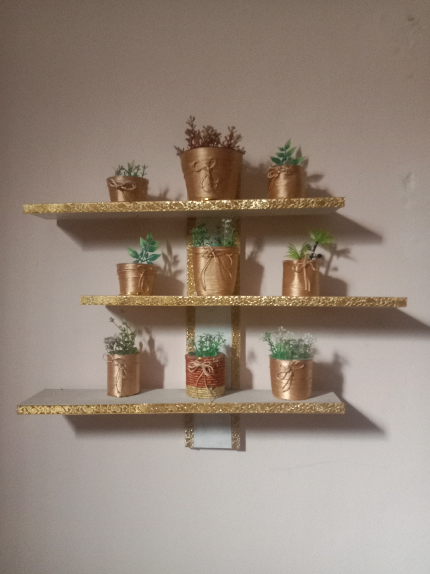 A wall-mounted shelf unit with three gold-trimmed shelves displays a collection of decorative potted plants. The shelves are staggered and extend outwards from a central vertical support. Each pot appears to be made of a gold-colored material and is adorned with twine or ribbon. The plants themselves are artificial, varying in type and color, with some featuring green foliage and others a more muted, dried appearance. The background is a plain, light-colored wall, suggesting an indoor setting. The image does not contain any elements that would allow for localization within the city of Kutosari, Indonesia.