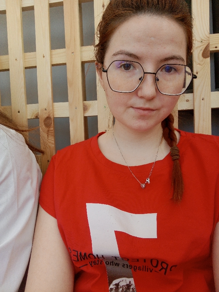 A close-up image depicts a young adult female, positioned from the chest up, looking directly at the camera with a neutral expression. She has fair skin and reddish-brown hair styled with a visible braid on her right side. She wears black, geometric-shaped eyeglasses. Her attire consists of a red short-sleeved t-shirt with a prominent white graphic on the front, beneath which inverted text, partially legible as "HOME" and "who stays," and a blurry monochrome image are printed. A thin, silver-colored necklace with a heart-shaped pink pendant and a smaller silver musical note pendant is worn around her neck. To the far left, a portion of a second individual, wearing a white shirt, is partially visible. The background consists of a light-colored wooden slat wall arranged in a grid pattern. The lighting is even, suggesting an indoor setting. The specific location within the city of Chaikovsky, Russia, cannot be determined from the visual evidence.