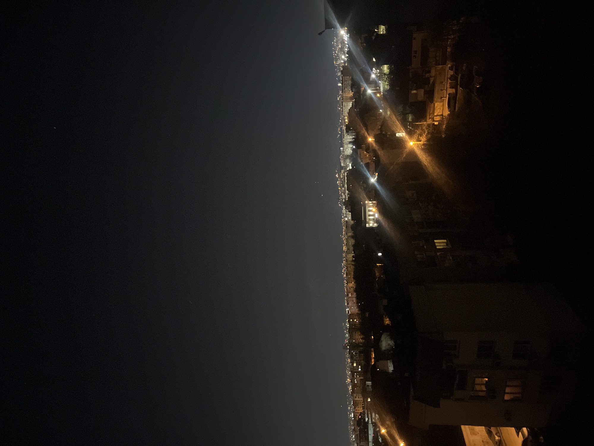The image depicts a nighttime cityscape, likely taken from an elevated position in Sofia, Bulgaria. The right side of the image shows buildings illuminated by internal and street lighting, with some appearing residential due to visible windows. The left side is dominated by a dark sky with few discernible stars. A horizontal stretch of lights likely represents the city's urban development stretching into the distance. There is no visible human activity or interaction. The environment appears to be urban with varying densities of buildings.