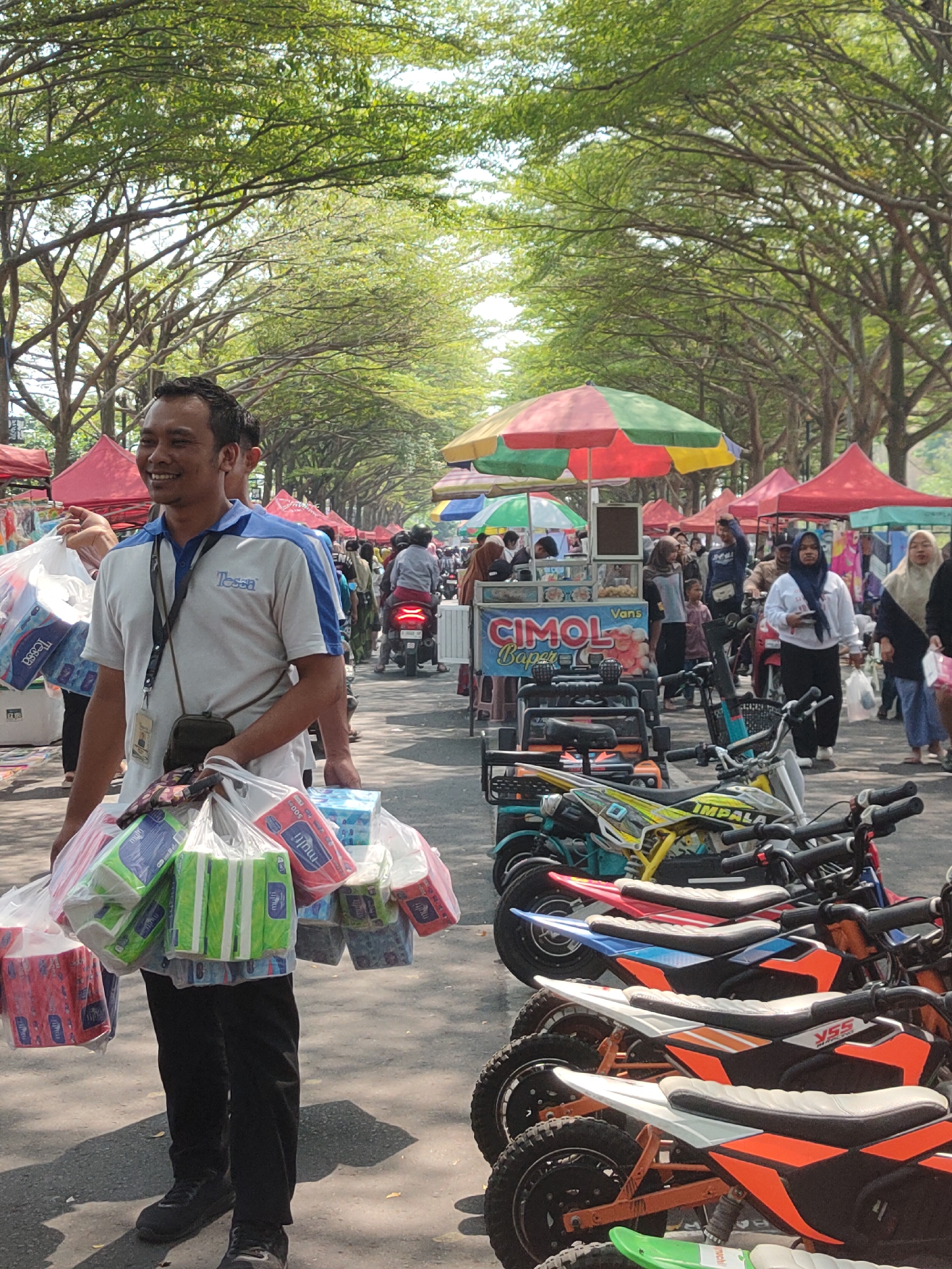 This image captures a bustling outdoor market scene, likely in Ngasem, Indonesia, characterized by rows of temporary stalls and a paved walkway lined with trees providing ample shade. The foreground is dominated by a smiling man wearing a blue and white polo shirt, carrying multiple large plastic bags filled with packaged tissue products. He is dressed in black pants and dark sneakers, with a small dark bag slung across his chest.
Behind him, the market unfolds with various stalls covered by brightly colored awnings, including red, yellow, and green tents. A food cart with the prominent signage "CIMOL" is visible, along with a display of what appear to be miniature motorcycles or scooters parked in a row, suggesting a mix of goods being sold. Several people are milling about, some on foot and others on motorcycles, indicating ongoing commercial activity and pedestrian traffic. The overall atmosphere suggests a lively and busy community market.