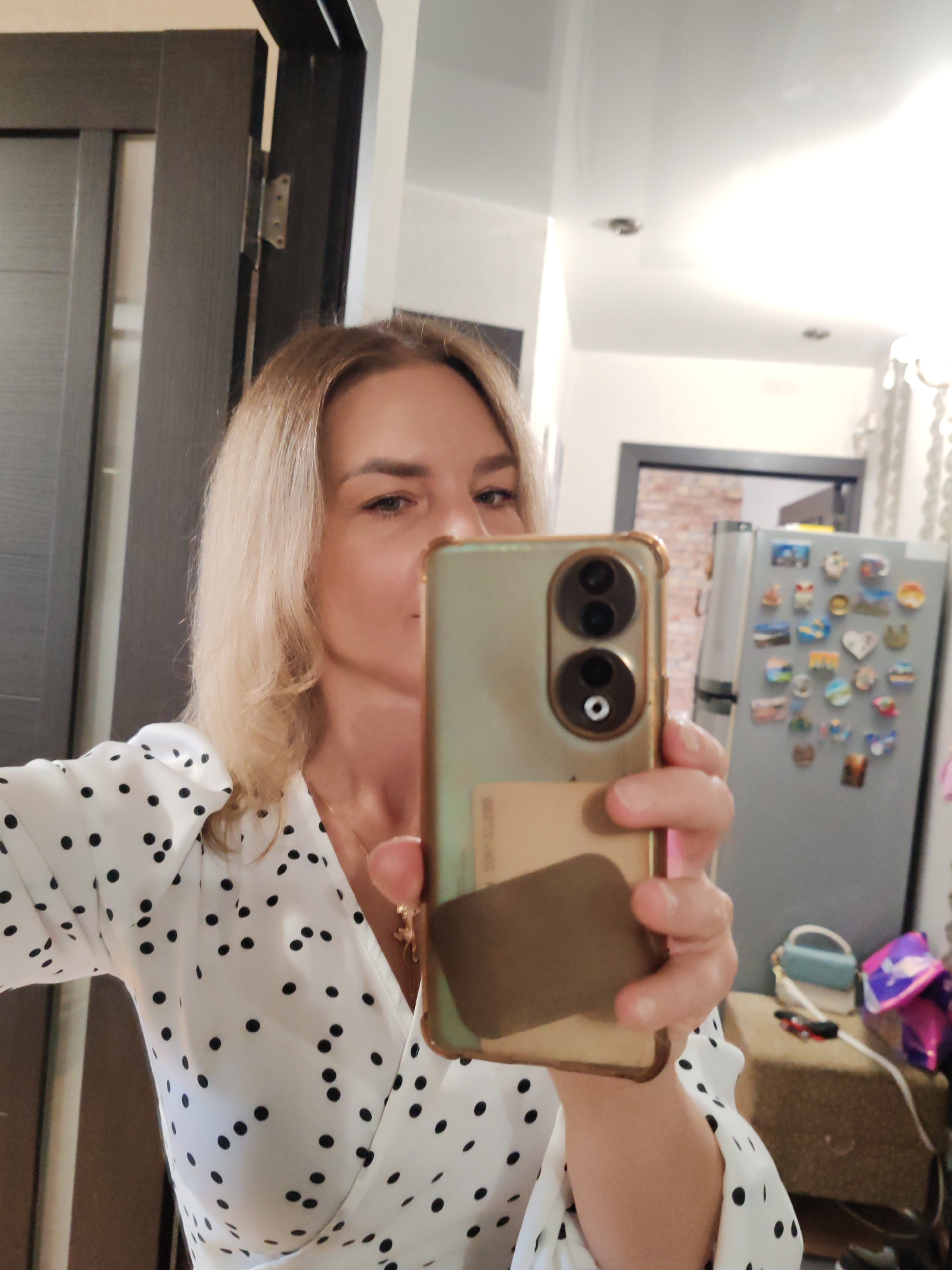 A blonde woman is depicted taking a mirror selfie indoors. She has light-colored hair, a neutral expression, and is wearing a white top with black polka dots. The woman holds a smartphone with a gold-colored case that has two prominent circular camera lenses on the back. A light brown card, possibly a credit card or ID, is tucked into the back of the phone case.
The background suggests a domestic setting. To the left, a dark wooden door and doorframe are visible, along with a metallic door hinge. On the right, a silver-colored refrigerator is covered with numerous colorful magnets of various shapes and sizes. Another dark doorframe is partially visible above and behind the refrigerator. The ceiling is white with recessed lighting fixtures, contributing to a brightly lit environment. Below the refrigerator, a beige upholstered bench or stool holds a small light blue and white purse and some shiny purple objects, possibly balloons or gift packaging. The image captures a casual moment, with no specific time of day indicated other than the artificial indoor lighting. No visible text is discernible in the image. The location context provided is Petropavlovsk-Kamchatsky, Russia.