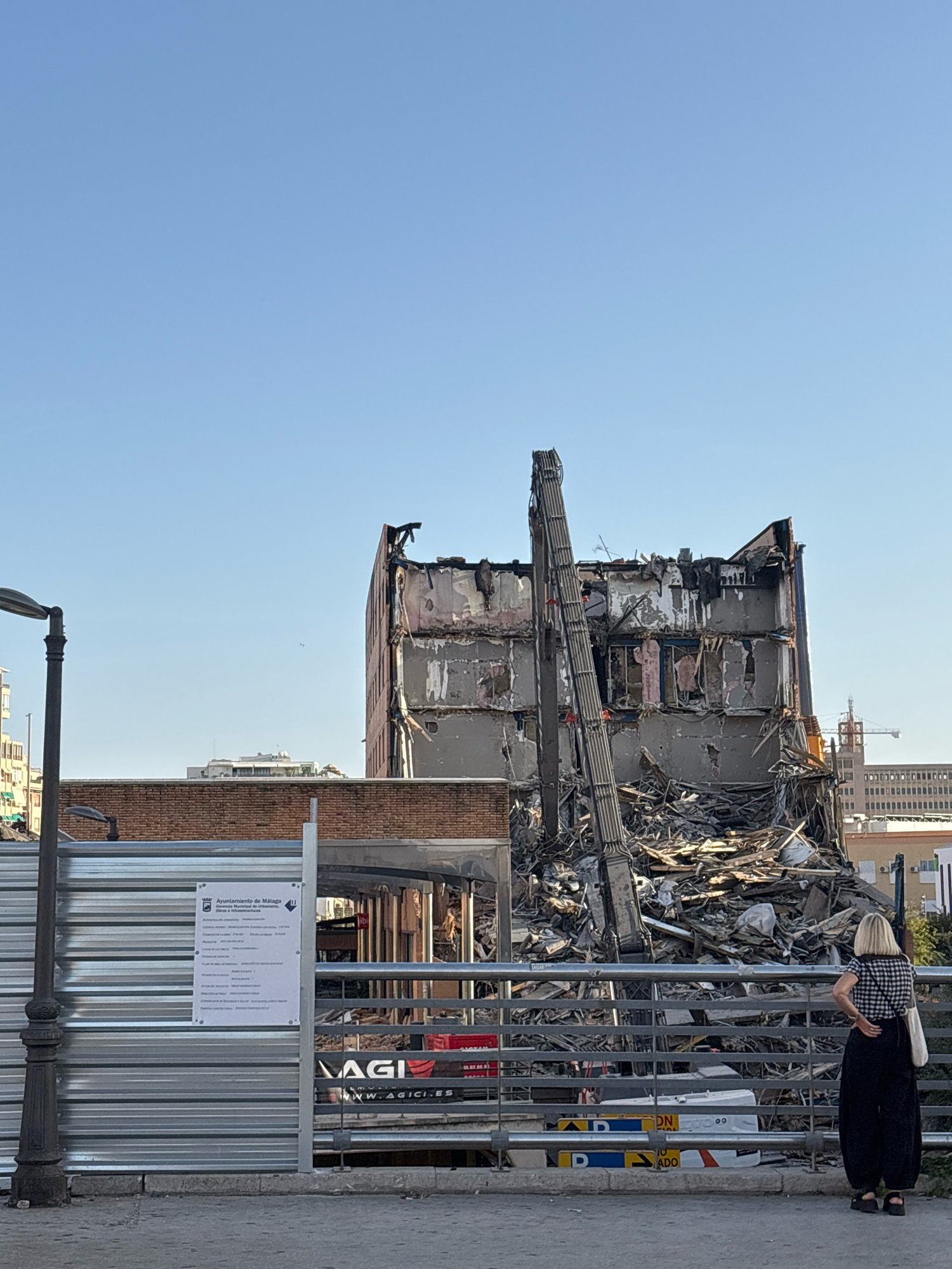 The image shows a woman standing and looking at a partially demolished building in Malaga, Spain. The building, which appears to be a multi-story structure, has had its upper floors removed, leaving a rubble-filled interior and exposed structural elements. A demolition excavator with a long arm is positioned against the side of the building. The scene is framed by a metal fence and a brick wall in the foreground. In the background, other city buildings and a clear blue sky are visible. A street lamp is present on the left side of the image. A sign is attached to the fence, which appears to be a notice regarding the demolition work, featuring the coat of arms of the City of Malaga.