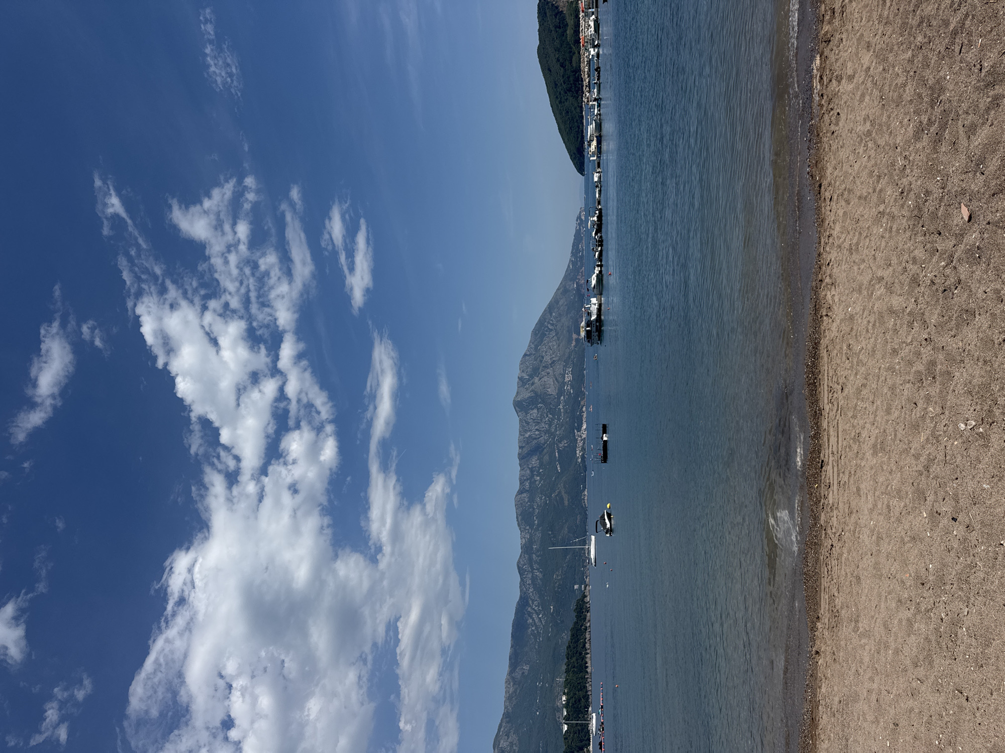 The image presents a vertically oriented view of a coastal scene, likely taken on a bright day, featuring a beach and seascape. The foreground shows a sandy beach, transitioning into the water's edge where gentle waves lap the shore. The water is a deep blue, which extends towards the middle ground of the image. Mountains are visible along the coastline, with the lower slopes covered in vegetation. Several boats are anchored in the water, and buildings dot the distant shoreline. The sky above is a mix of clear blue and scattered white clouds. This view is likely a panorama taken in the city of Budva, Montenegro.