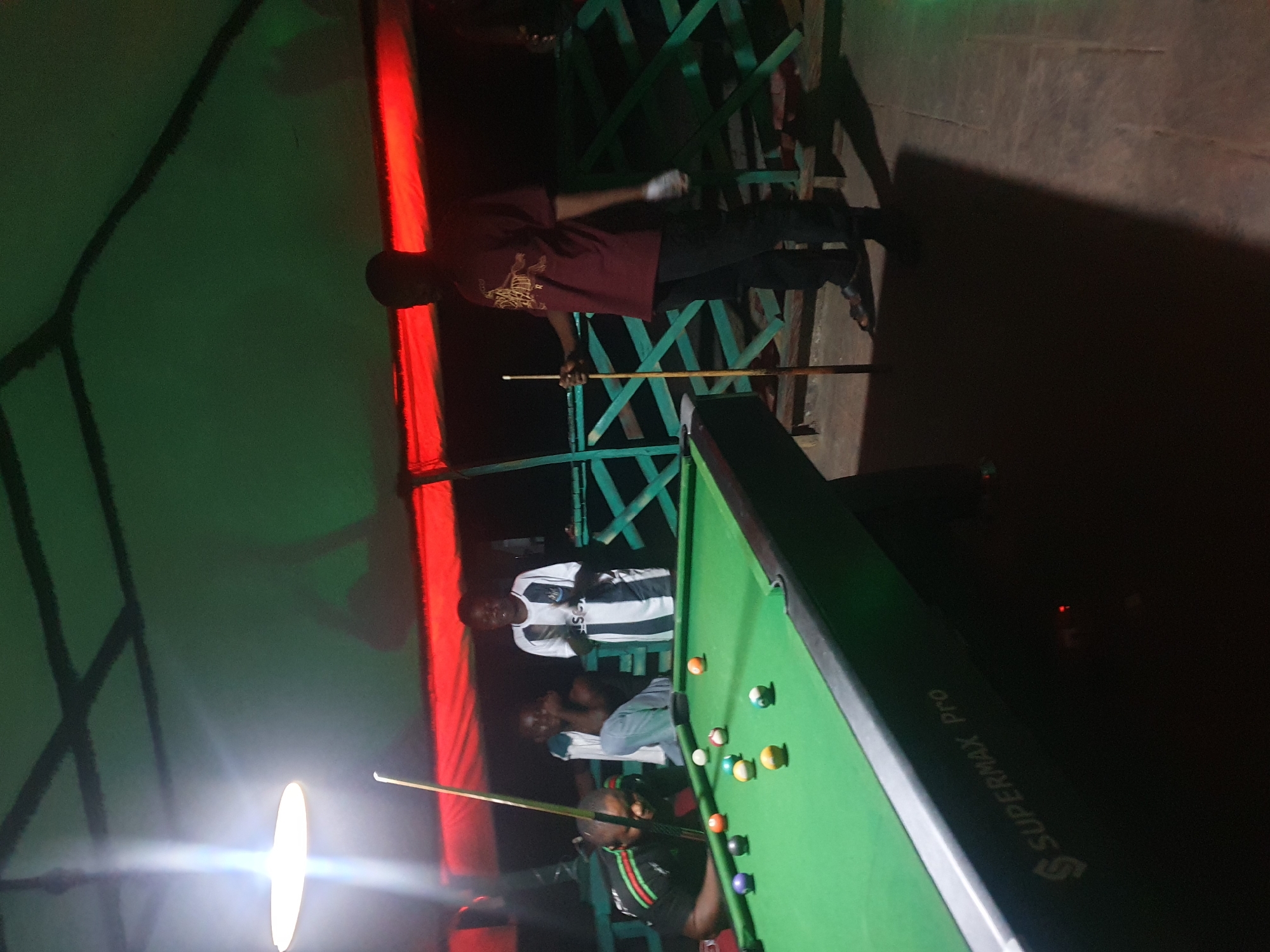 The image depicts a group of people playing pool at an outdoor establishment, possibly in Lokoja, Nigeria. The scene is dimly lit, with a large overhead light illuminating the pool table and a portion of the surrounding area. A bright red fabric or tarp is visible overhead, casting a reddish glow on the scene. The pool table itself is green, with several billiard balls scattered across its surface.
Several individuals are present. In the foreground on the left, a man wearing a black shirt with green and red trim is leaning over the pool table, appearing to be in the process of playing. To his right, another man in a white and black striped jersey is standing and watching. Behind them, two other individuals are seated, their faces partially obscured by darkness. On the right side of the image, a man in a maroon t-shirt with a graphic print and dark pants is holding a pool cue. He is standing near the edge of the pool table, facing away from the camera. The environment appears to be a casual outdoor setting, with green wooden fencing visible in the background. The words "SUPERMAX Pro" are visible on the side of the pool table, suggesting the brand of the equipment.