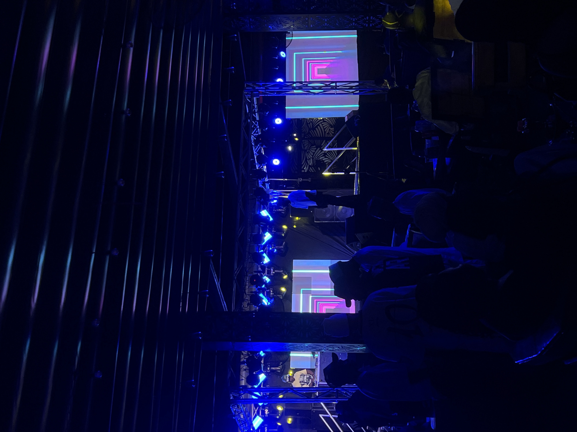 The image depicts a dimly lit indoor event, likely a concert or performance, taking place in Howayi, Nigeria. A vertical metal corrugated wall is visible on the left. Multiple people are seated and standing, mostly in silhouette. A stage is constructed with lighting and a large screen displaying colorful geometric patterns. A performer is visible on stage in the center. The overall color palette is dominated by shades of blue and purple, emanating from the lighting fixtures and the screen.