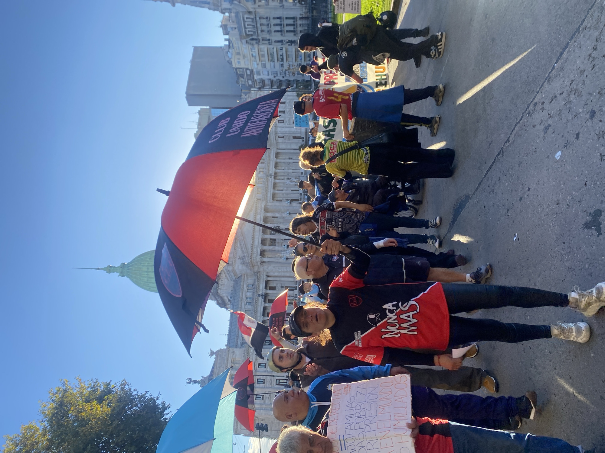 An outdoor scene captures a public gathering, likely a demonstration or protest, under a bright, clear blue sky in Buenos Aires, Argentina. The prominent dome of the Argentine National Congress building is visible in the background, anchoring the location.
Numerous people are present, forming a crowd that extends into the distance. Many are holding open umbrellas in various colors, including red and black, light blue, and red, possibly for shade from the strong sunlight. Some participants are holding banners or signs. The time of day appears to be sunny daytime, with harsh shadows indicating strong sunlight.
Visible text on an umbrella reads "CLUB UNIDO ANTIFASCISTA." Another umbrella shows "CLUB UNIDO." A person in the foreground wears a black and red shirt with "NUNCA MAS" (Never Again) emblazoned on it, a significant slogan in Argentina related to human rights. A man in a blue jacket holds a white sign that reads:
"CONTRA LA DERECHA"
"TRABAJO FAMILIAS"
"POBREZA"
"REPRESION"
"EN EL MUNDO"
"HUMILLADOS"
Other partially visible texts include "LA PATRI DEFIEND" (likely "La Patria se defiende" - The Homeland is defended) on a banner, and "difacil" on a person's yellow and green striped shirt. The scene conveys a sense of organized public dissent or advocacy.