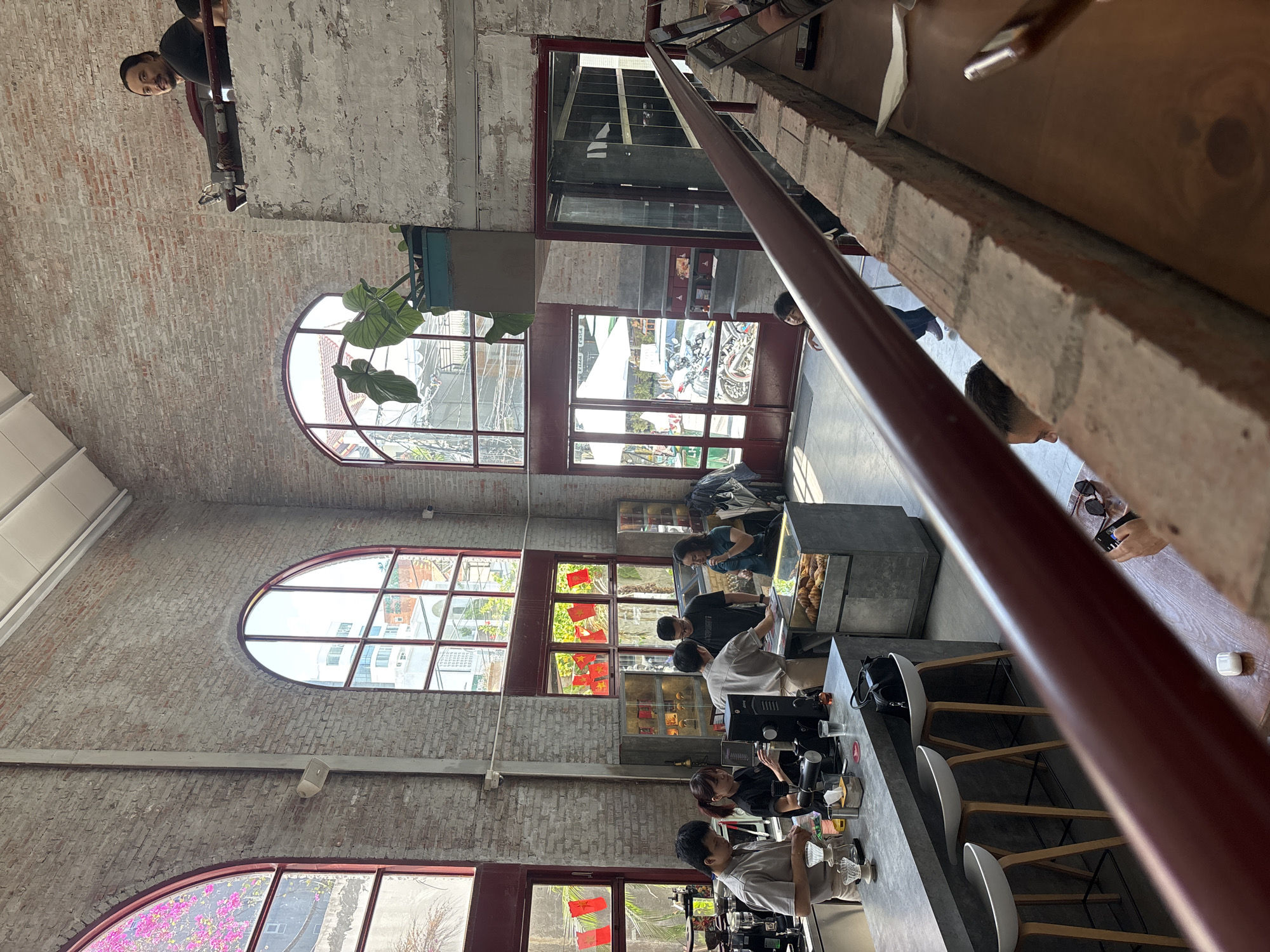 This image depicts the interior of a bustling cafe in Da Nang, Vietnam, characterized by exposed brick walls, large arched windows, and industrial-style furnishings. In the foreground, a dark red railing runs diagonally across the frame, obscuring a portion of a wooden table with a pair of sunglasses and a phone on it.
To the left, a bar area is staffed by several baristas. One male barista, wearing a light brown uniform, is preparing coffee, while a female barista in a black uniform assists. Behind them, a display case filled with croissants is visible. Other customers stand near the counter, engaged in ordering or observing the preparation.
In the background, large arched windows offer views of the city, with trees and buildings visible outside. One window is adorned with bright pink flowers. On a higher level of the cafe, a man with dark hair is seated, looking towards the camera. The overall ambiance suggests a lively and trendy establishment. The architecture and decor imply a blend of industrial and modern aesthetics.
