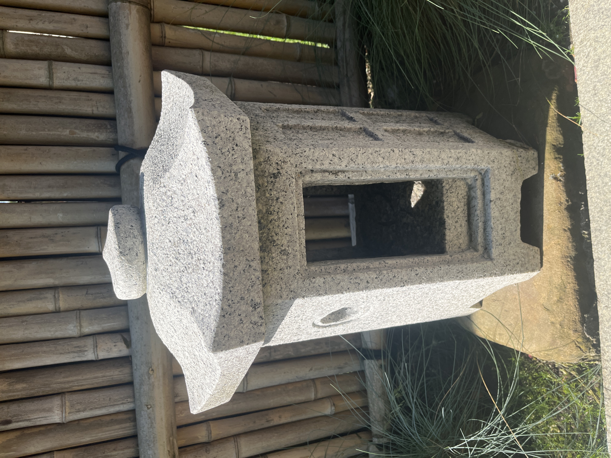 An outdoor, eye-level, full shot captures a Japanese stone lantern positioned in front of a bamboo fence. The lantern, made of speckled grey granite, is hollow with an open front and a circular opening on its left side. It rests on a flat, brownish rock. Surrounding the lantern is a patch of green grass and thin, wispy green foliage. The bamboo fence behind the lantern is constructed from vertical, light-brown bamboo stalks. Sunlight casts shadows on the lantern, indicating it is a sunny day. There are no people or animals visible in the scene. The image depicts a peaceful, garden-like setting.