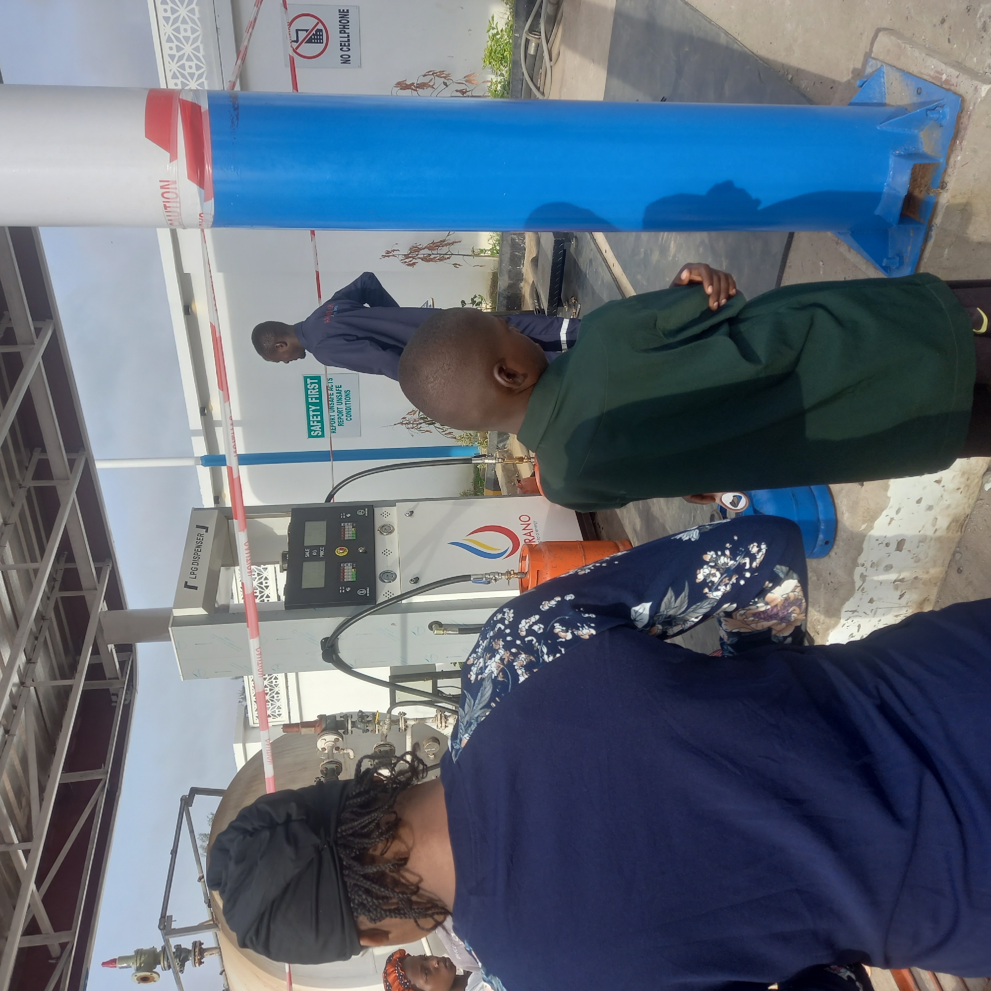 The scene captured at a gas station in Barden Yana, Nigeria, shows a gas dispenser with signage indicating it's an LPG (Liquefied Petroleum Gas) dispenser. The pump's display panel is visible. A sign is also posted with the text "SAFETY FIRST: REPORT UNSAFE ACTS, REPORT UNSAFE CONDITIONS." Another sign warns, "NO CELLPHONE."
Several people are present. A man in a dark blue uniform stands near the gas pump, and a child in a green shirt is also in the frame. A woman wearing a navy blue top with a floral pattern on the sleeves and a head covering is present.
The environment appears to be a typical gas station setting with metal structures and poles painted in white, blue, and red. Orange gas tanks sit nearby. Caution tape is wrapped around the pump.
