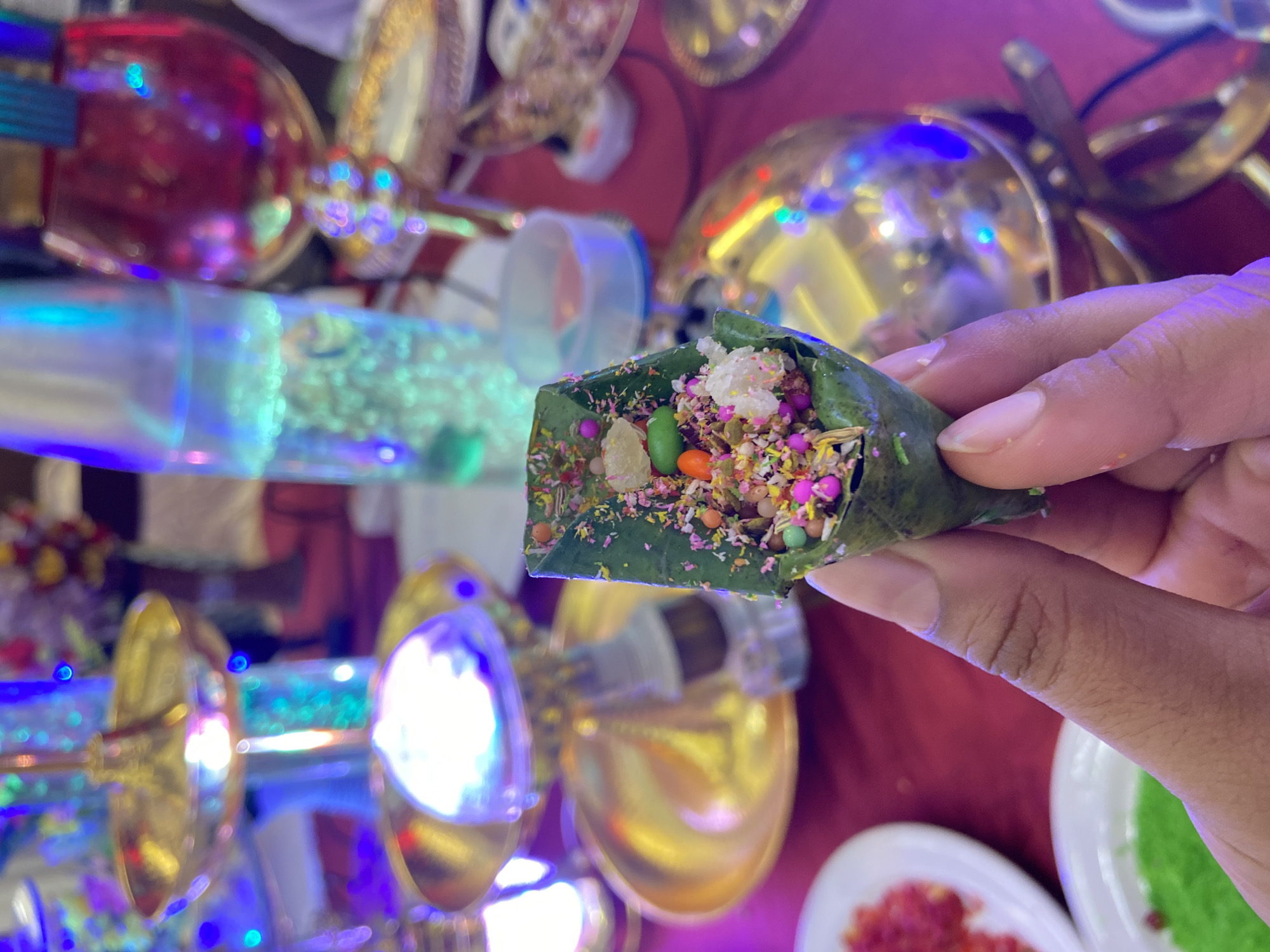 A hand holds a betel leaf paan filled with colorful ingredients against a backdrop of decorations. The filled paan appears to contain various candies and sweet seasonings. The person holding the paan is of brown skin complexion, with short fingernails. The background is filled with decorative elements, including golden objects, lights, and what appears to be a maroon tablecloth. The image gives the impression of being at an event or celebration where paan is being served. It is possible this image was taken in Dhaka, Bangladesh given the local culinary tradition.