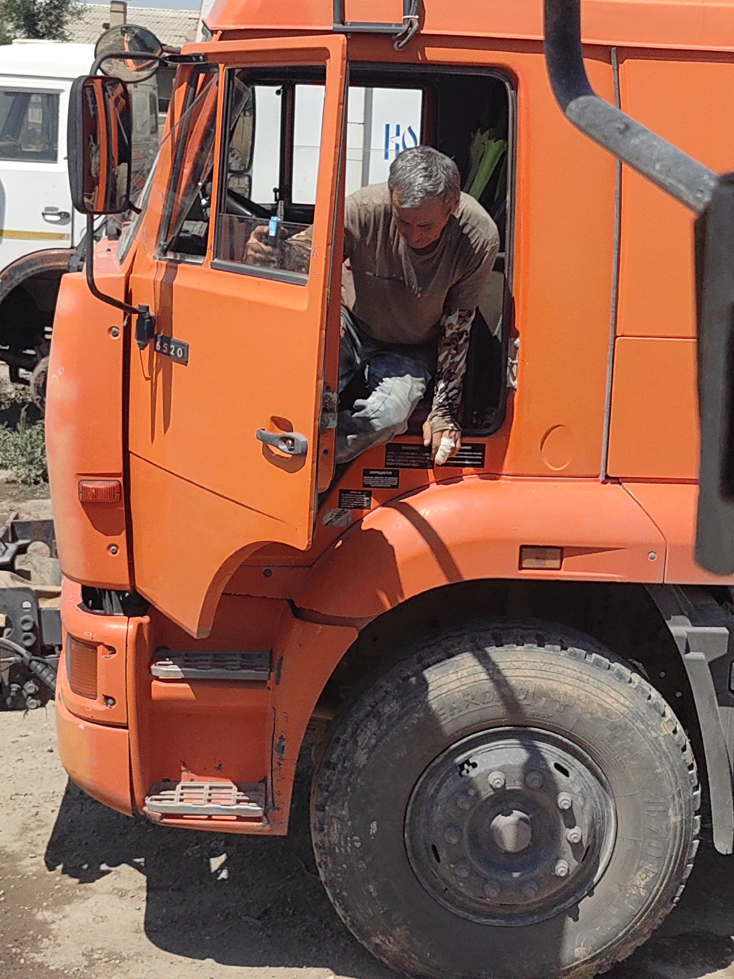 In Shymkent, Kazakhstan, a man with short gray hair is leaning out of the open driver's side door of an orange truck. He is wearing a brown t-shirt and camouflage-patterned sleeves on his arms. His right hand, which has a bandage on it, is resting on the truck's frame. The truck has the number "6520" on its side, above the door handle. The environment appears to be outdoors, with a dusty ground and some vegetation visible. Another white truck is partially visible in the background to the left. The image captures a close-up of the truck's cab and the man within it.