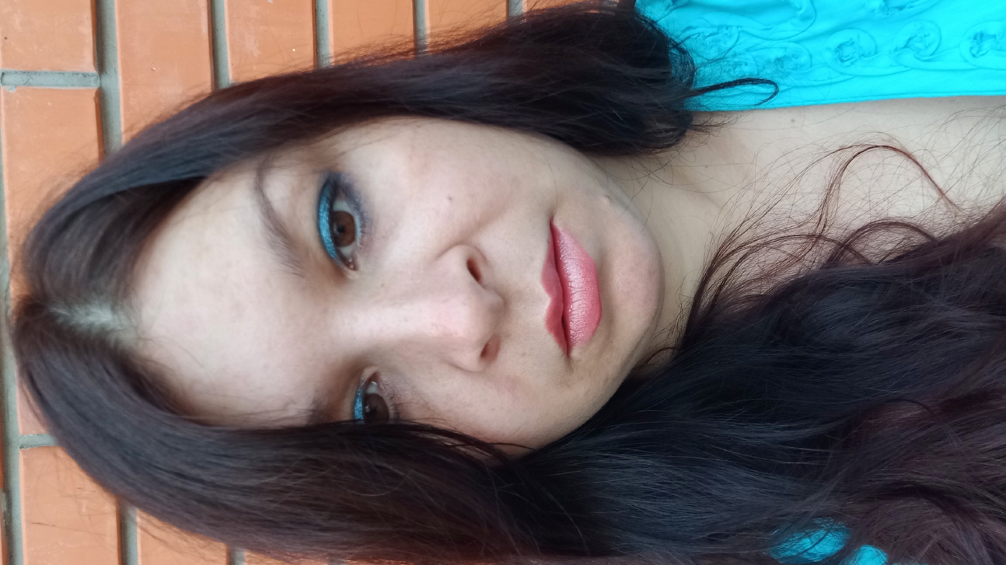 A medium-shot photograph captures a woman's face and upper torso against an orange brick wall. The woman has dark brown hair, which falls to her shoulders, and is wearing blue eye shadow and pink lipstick. Her expression is neutral. The brick wall, a common urban feature, is likely located in Kropyvnytskyi, Ukraine, given the prompt's context. The lighting suggests it may be daytime.
