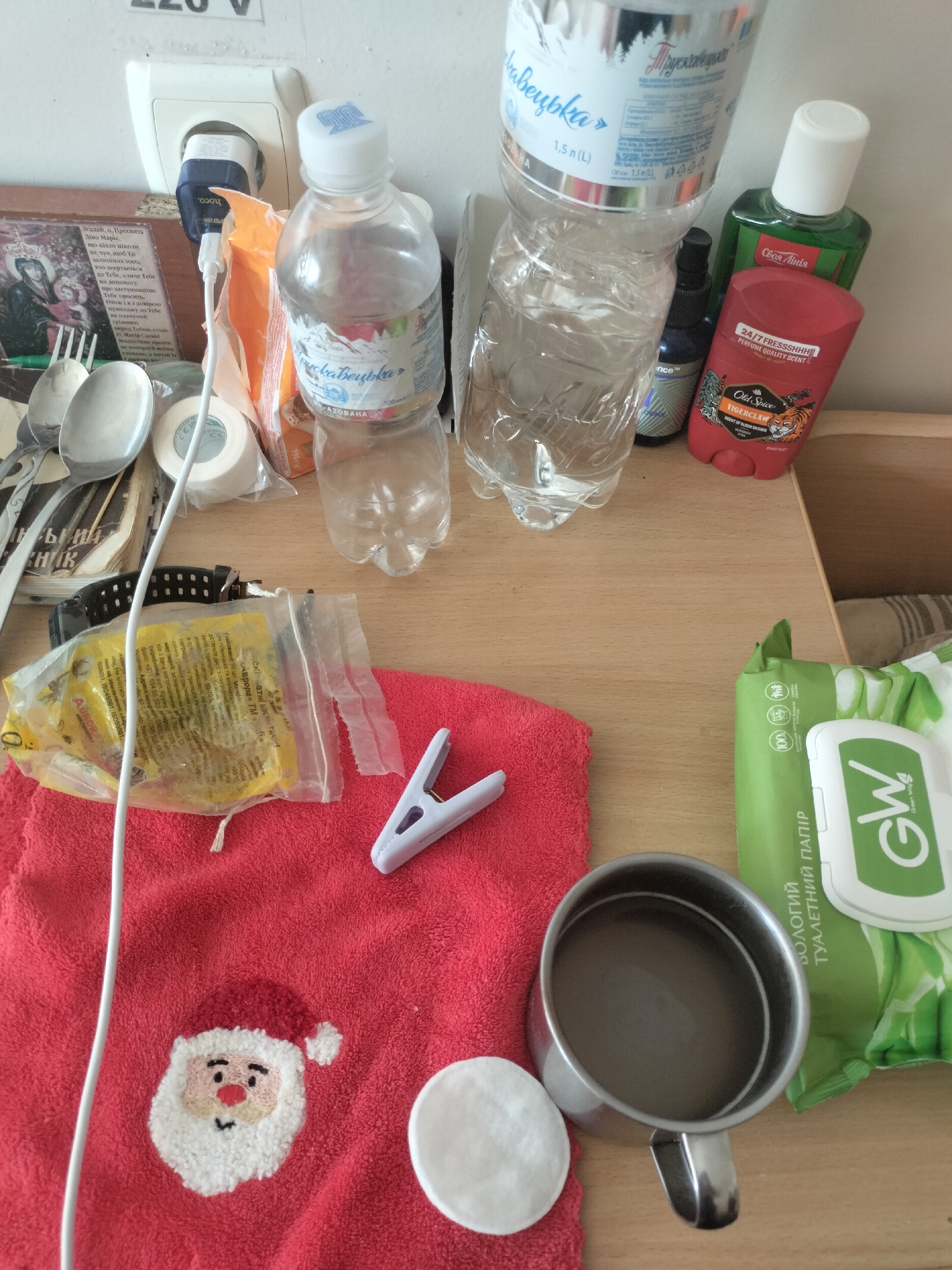 The image depicts a cluttered wooden table with various personal items. In the upper left corner, an electrical outlet is visible with a plug inserted. To the right of the outlet are two plastic bottles of water. A green bottle of what appears to be aftershave and a red deodorant stick are placed next to the water bottles. In the center of the image, a red towel with an embroidered Santa Claus face is laid out. On the towel, there is a white plastic clothespin and a small yellow plastic bag containing some items. To the right of the towel, a package of wet wipes with the brand "GW" is visible. A stainless steel mug containing a dark liquid is also present on the table. A white cotton pad rests on the towel near the mug. In the background, a framed picture is partially visible, and a watch with a black strap is lying on the table. The overall environment suggests a personal space, possibly a room or a desk area. The location is identified as Unknown, Unknown.