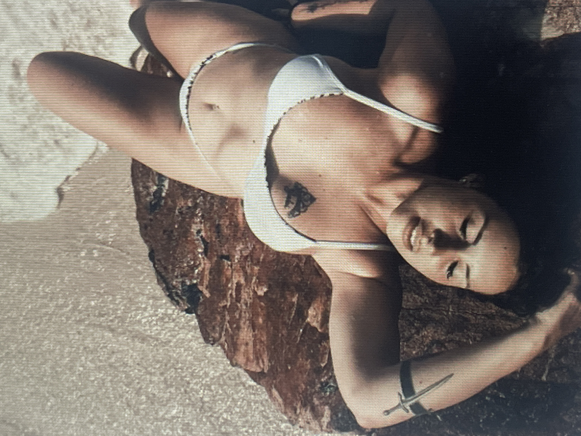 The media file displays a person lying on a rocky surface near the water. The person is wearing a white bikini and has a tattoo on their chest. There is also a decorative band with a small sword on their upper arm. The scene appears to be outdoors, possibly a beach or coastal area, with sand and water visible. The lighting suggests it could be daytime, but the overall image quality is grainy, making it difficult to ascertain specific details about the weather or exact time of day. The person's facial expression is relaxed, with their eyes closed and a slight smile. The image is oriented such that the person is lying on their back.