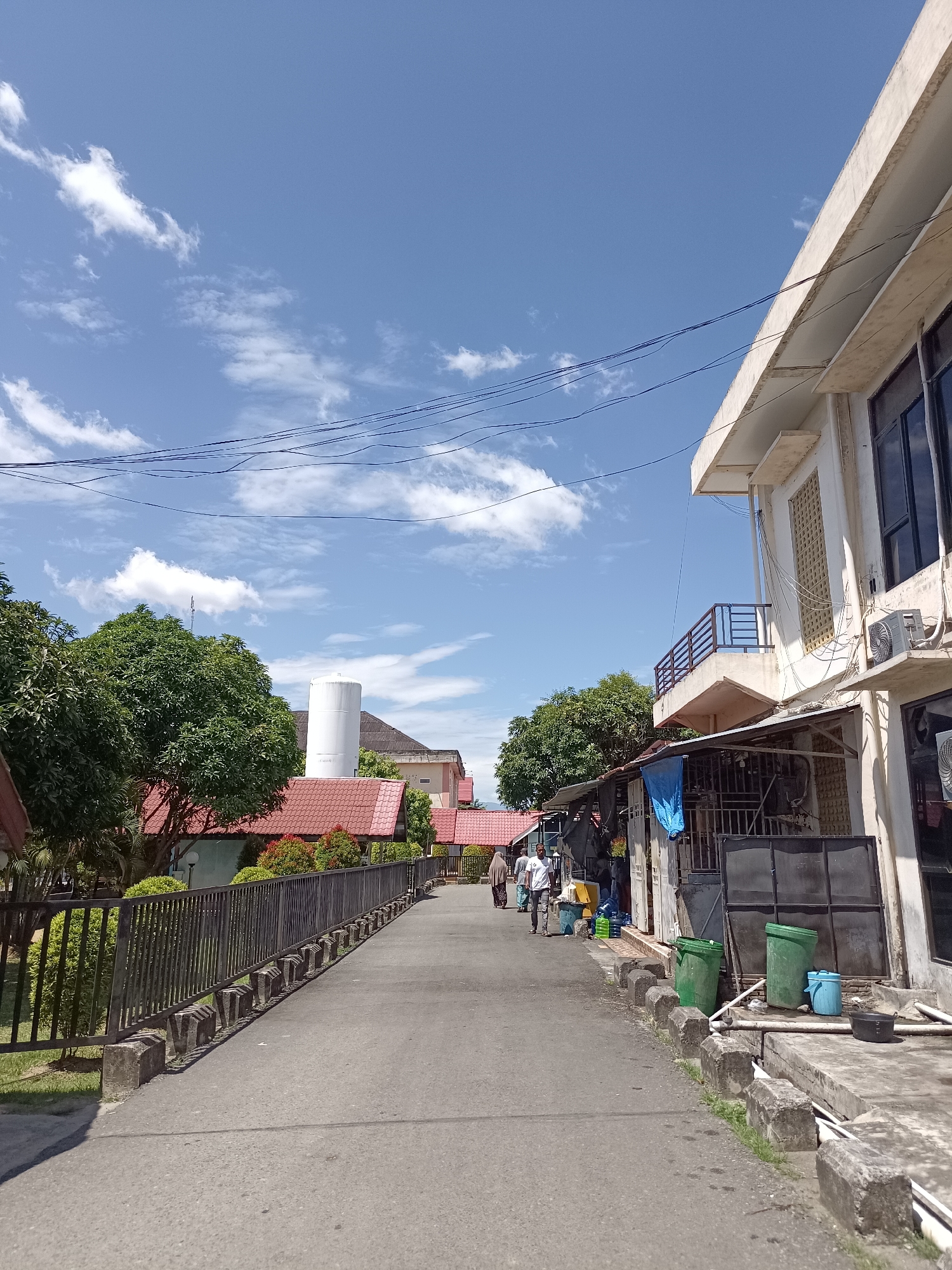 The image depicts a street scene in Mesjid Reuntoh, Indonesia, under a bright blue sky with scattered white clouds. On the right side of the paved street, a two-story building with white walls and dark windows is visible, featuring a balcony on the upper level. Below the balcony, a small storefront with a corrugated metal roof and a blue tarp is present. Various containers, including green trash bins and blue water jugs, are situated near the storefront.
To the left of the street, a metal fence with concrete posts lines the pathway, separating it from a grassy area and lush green trees. Beyond the fence, buildings with red tile roofs are visible, along with a tall white cylindrical structure, possibly a water tower, further in the background. Several individuals are walking along the street in the distance. The overall impression is a sunny day in a semi-urban or residential area of the city.