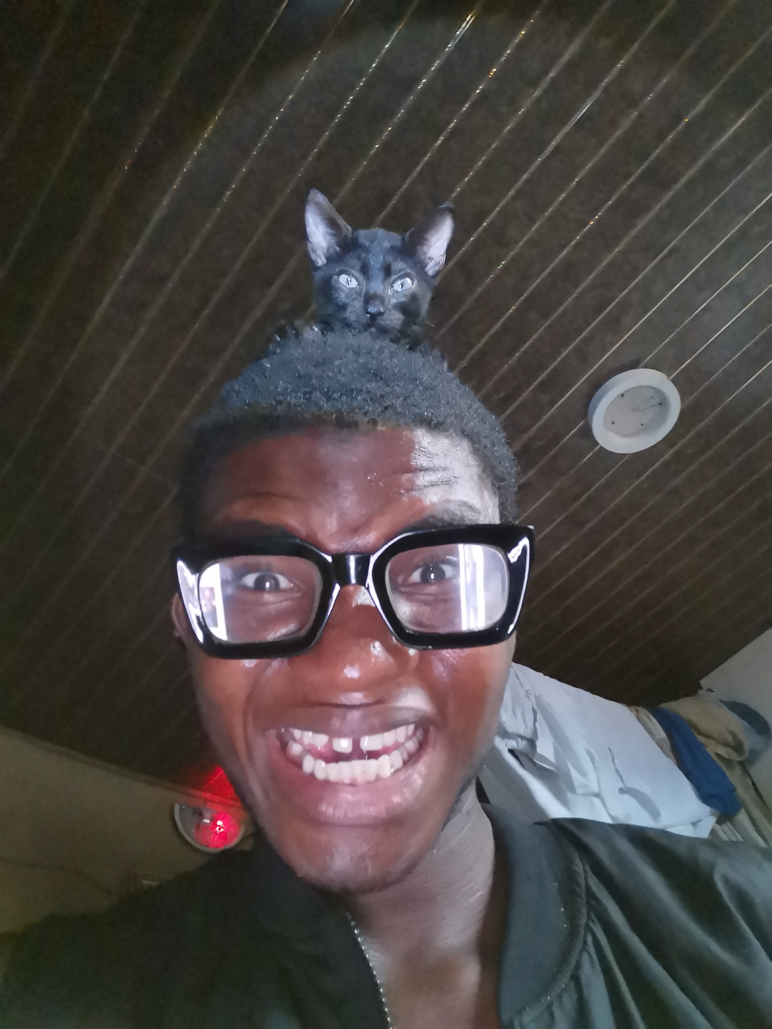 A man is posing for a selfie with a small black cat on top of his head. The man is wearing large, black-framed glasses and has a wide smile. He has dark skin and short, curly black hair. He is wearing a dark jacket. The cat is small with blue eyes. The background appears to be an indoor setting with a ceiling of brown wooden planks. In the background, a white light fixture is visible on the ceiling. There is also a small red light and some hanging clothing or bedding. Based on the context of Lagos, Nigeria, this likely takes place inside a residence, although the exact location cannot be determined from the image.