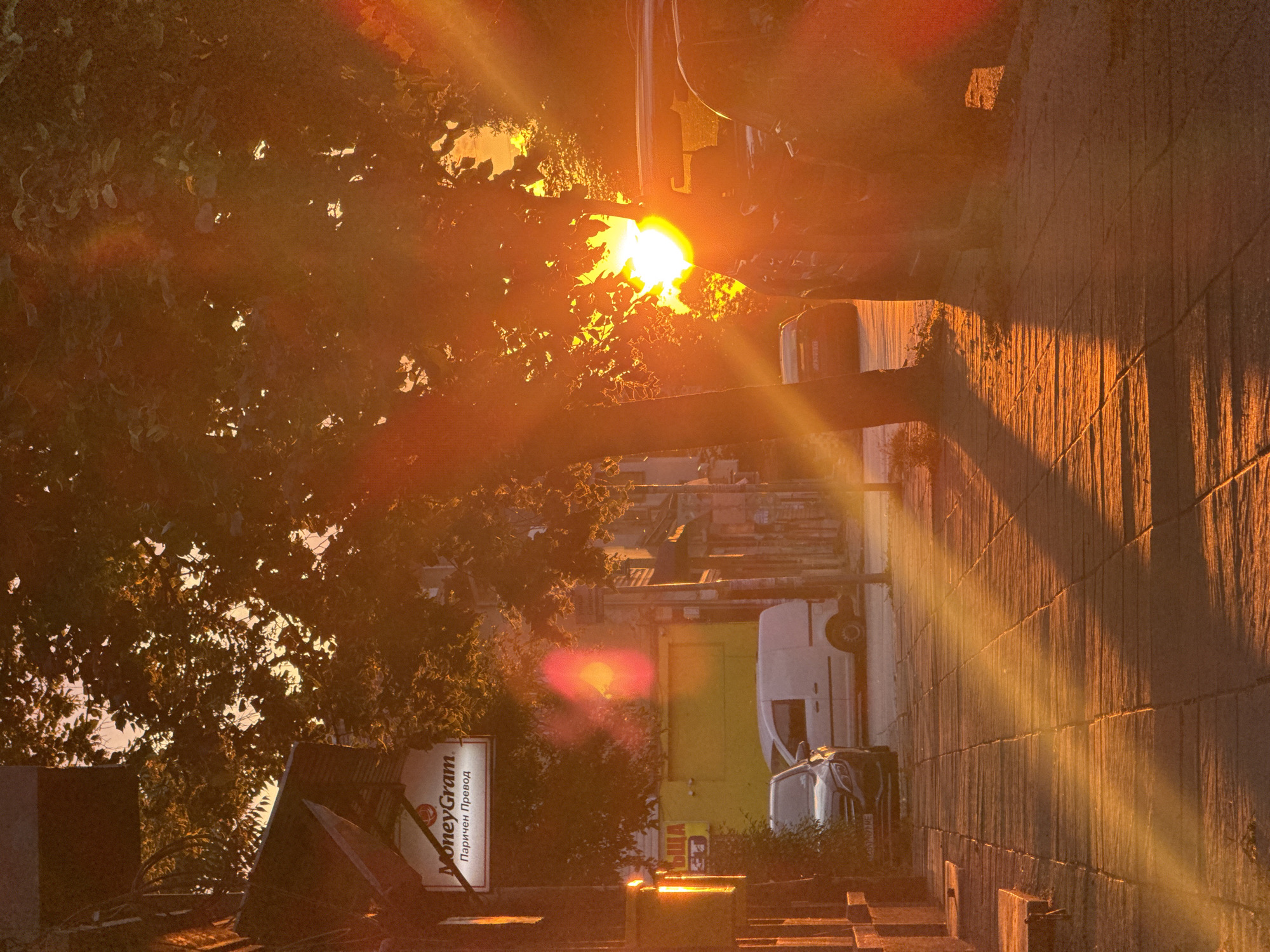 The image captures an urban street scene illuminated by intense, low-angle sunlight, creating strong lens flares and elongated shadows. The location is identified as a street in Sofia, Bulgaria, evidenced by a prominent "MoneyGram" sign featuring the English text "MoneyGram" and the Bulgarian text "Паричен Превод" (Money Transfer).
In the foreground, a paved sidewalk extends from the lower left, textured by the warm light and shadows. Two tree trunks rise from the sidewalk on the left. Along the right side of the street, several vehicles are parked. A dark-colored sedan, bearing design characteristics consistent with a Mercedes-Benz model, is partially visible on the left side of the street, while a white panel van is parked further down.
The midground features several buildings. The "MoneyGram" sign, white with red and black lettering, is affixed to a building on the left. Adjacent to it, another building features a uniformly yellow facade, appearing to be closed. Dense green foliage from multiple trees partially obscures the buildings and the sky, with leaves brightly backlit by the sun. A brilliant, circular sunburst with distinct rays and multiple orange-red lens flares is positioned in the right-center, partially obscured by tree foliage and a parked vehicle. A partially visible street sign, featuring Bulgarian script, is also present on a pole to the right.
The background shows glimpses of a hazy, light orange sky through the tree canopy, and outlines of distant urban structures. No people are visible within the scene. The dominant colors are warm oranges, yellows, and deep browns in the shadowed areas.