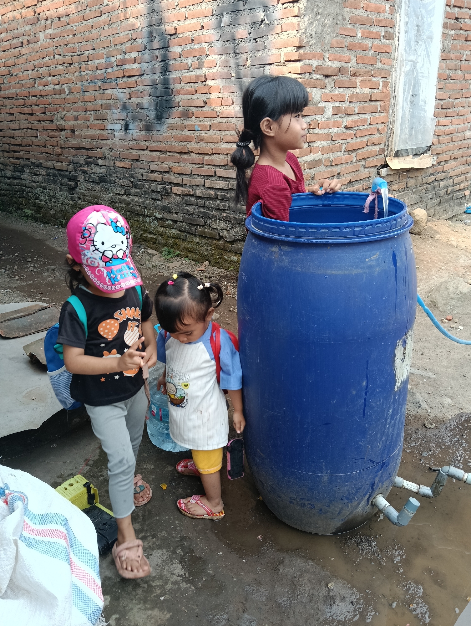 Three young girls are gathered around a large blue plastic barrel filled with water. The girl on the far right, wearing a red shirt, is leaning over the barrel, appearing to interact with the water or something inside. The girl in the middle, wearing a white and blue shirt and yellow shorts, is looking down at the barrel, possibly reaching into it. The girl on the left, wearing a black t-shirt with a cartoon print and a pink Hello Kitty baseball cap, stands slightly apart, looking to her right. All three girls are wearing backpacks, suggesting they are on their way to or from school, or engaged in an activity that requires them. The barrel has a faucet attached near the bottom, from which a blue hose extends. The ground is muddy and wet, indicating recent rain or spillage. The scene is set against a rough brick wall. The image captures a moment of everyday life, possibly related to water collection or a communal water source, in a residential area of Tasikmalaya, Indonesia, likely in a modest urban or semi-urban setting characterized by its brick architecture and unpaved ground.