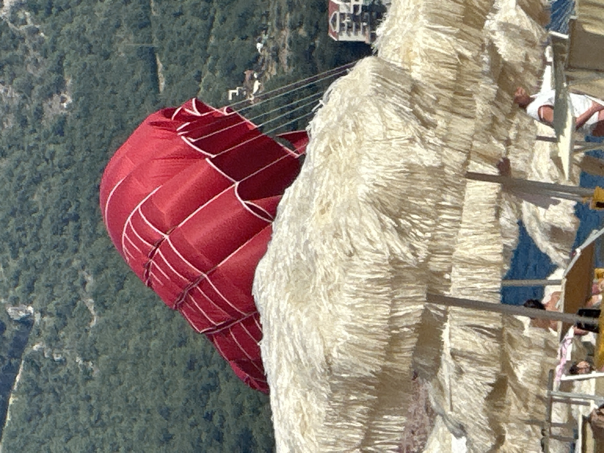 In Boreti, Montenegro, a red hot air balloon is partially visible against a backdrop of lush green hills. The balloon, adorned with white stitching, appears to be descending or preparing for landing. In the foreground, a row of thatched beach umbrellas casts shadows over the sand, with a few beach chairs and obscured figures suggesting a relaxed atmosphere. The sea is visible in the distance, a calm blue expanse. The scene captures a moment of leisure and adventure, with the balloon adding an element of unexpected spectacle to the tranquil beach setting.