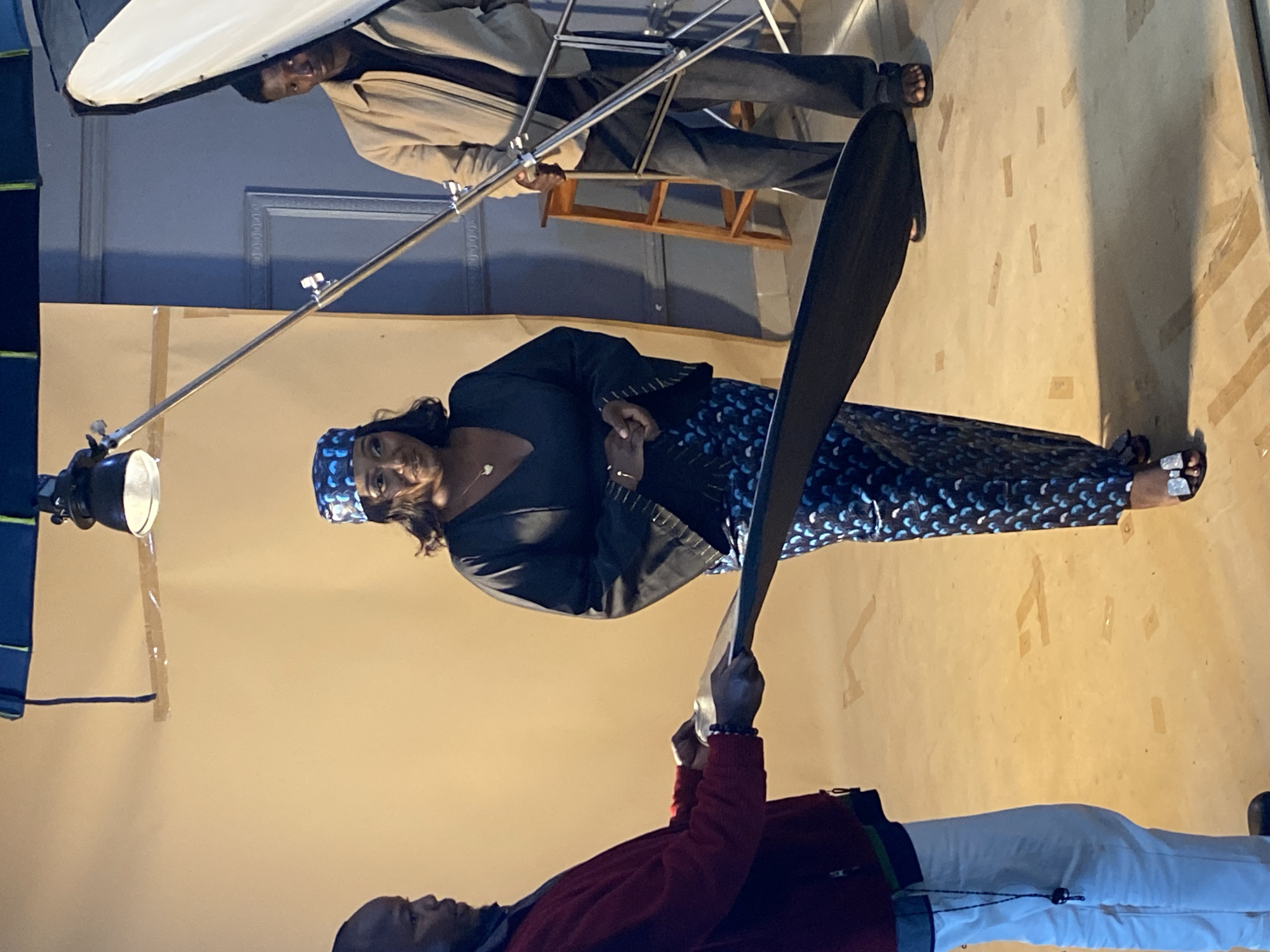 In Bukuru, Nigeria, a woman stands in the center of a studio, wearing a patterned headscarf, a V-neck black top with embellished sleeves, and a matching patterned skirt. She has her hands clasped in front of her. To her left, a man in a red jacket and white pants holds a large black reflector, looking towards her. To her right, a man in a light brown jacket and dark pants stands near a wooden stool and a lighting rig. The background is a plain beige wall, with a dark blue wall and a door visible on the right. The floor is a light-colored surface with various strips of tape. A studio light is suspended from the ceiling.
