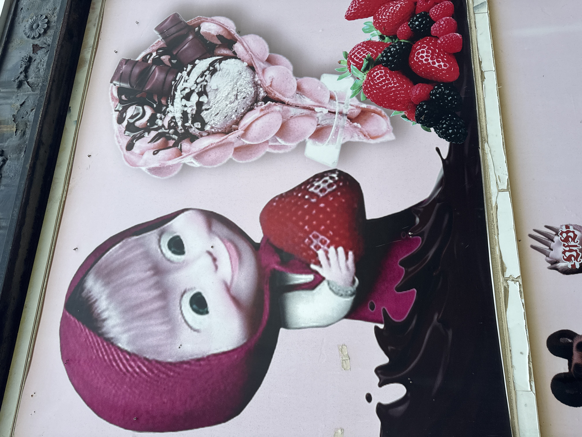 An outdoor advertisement displays a cartoon of a young girl in a pink headscarf holding a large strawberry, with a portion of chocolate dripping below her. To the right of the girl is an ice cream cone made of bubble waffle with chocolate sauce and candy bars. A pile of strawberries, blackberries, and raspberries is in the lower right corner of the advertisement. The advertisement is framed by a tarnished metal border at the top and a damaged white frame at the bottom. The advertisement appears to be in a state of disrepair, with peeling paint and tape on its surface. The background of the advertisement is a pale pink. There are no people present in the image.