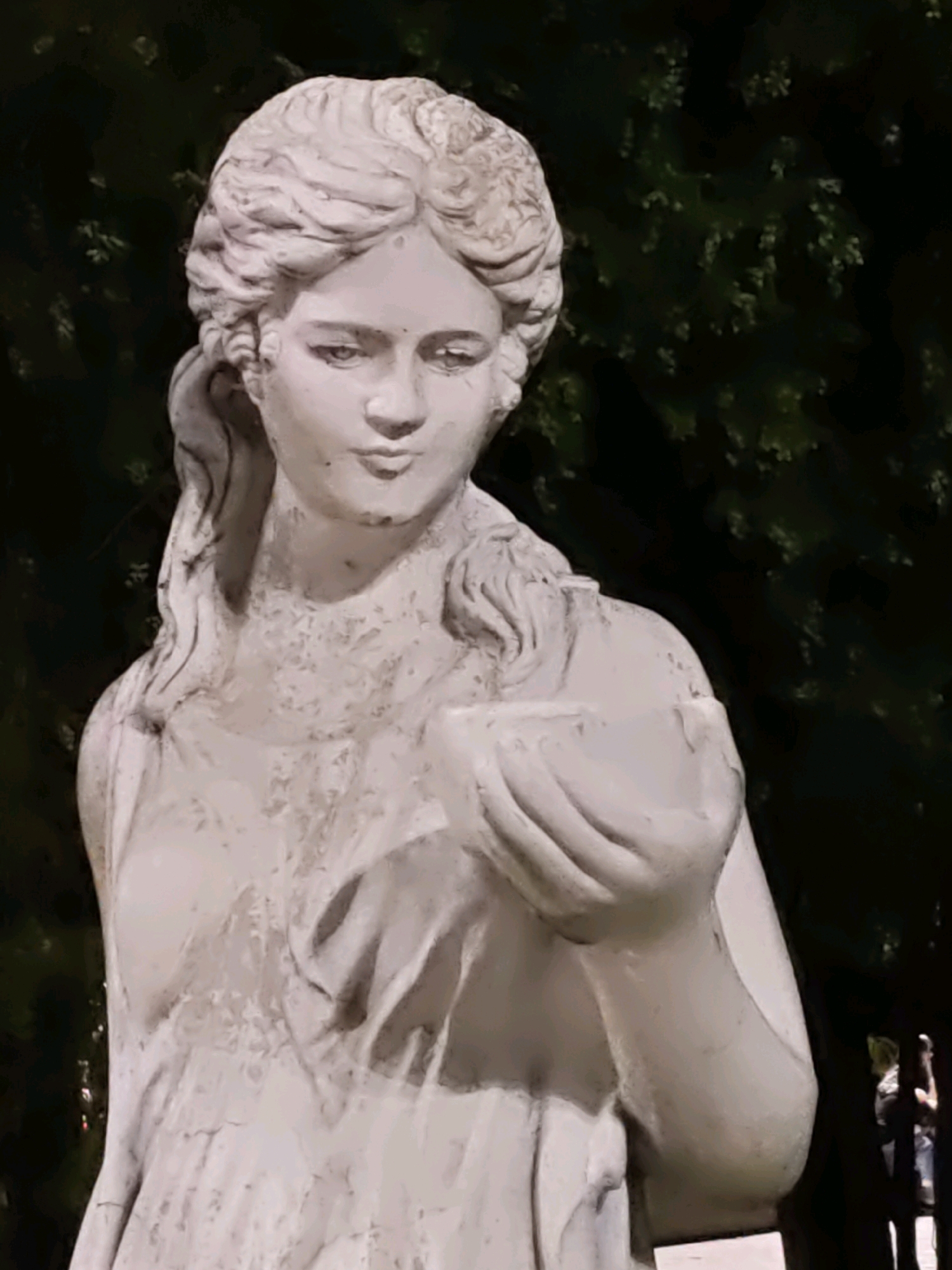 A white stone statue of a woman holding a bowl stands in front of a dark green, leafy background. The statue depicts the woman from the chest up, with her head turned slightly to her left. Her hair is styled in an updo with curls framing her face. She wears a draped garment. The statue is weathered, with visible cracks and imperfections. In the background, out of focus, a dark shadow of a person can be seen holding a phone or camera. The lighting suggests it is either early morning or late evening. This statue is located in the Park of Culture and Leisure named after Maxim Gorky in Rostov-on-Don, Russia.