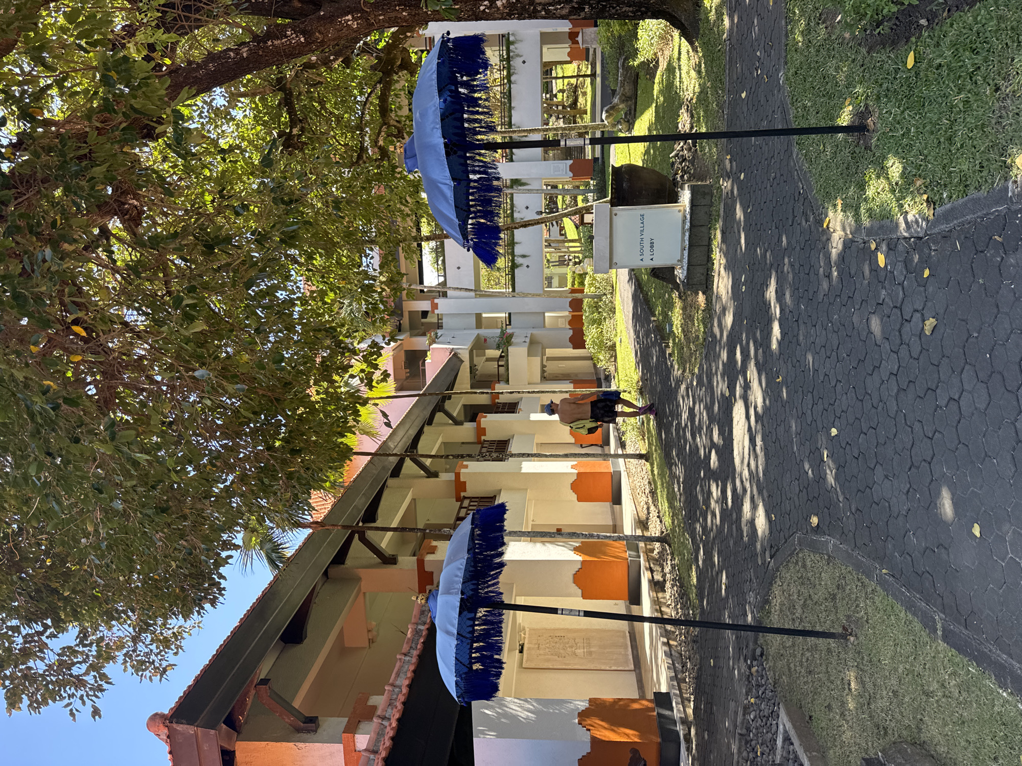 The image depicts an outdoor scene in Kuta Selatan, Indonesia, featuring a paved walkway lined with two light blue ornamental umbrellas with dark blue fringe. A multi-story building with white pillars and orange accents runs parallel to the walkway. A shirtless man wearing shorts, a blue hat, and a yellow backpack walks on the pathway. The area is shaded by trees, creating dappled sunlight patterns on the walkway. A sign indicates the location is near a South Village lobby.
